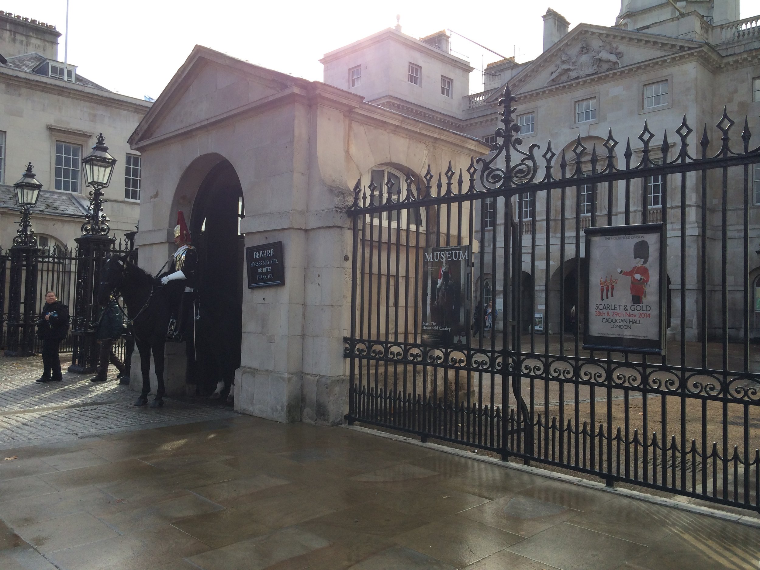 View of a historic building, possibly a museum, with a gated entrance and a guard in the form of a mounted soldier. There are signs indicating it is a museum, and the surroundings include old-fashioned street lamps and a few people standing near the 