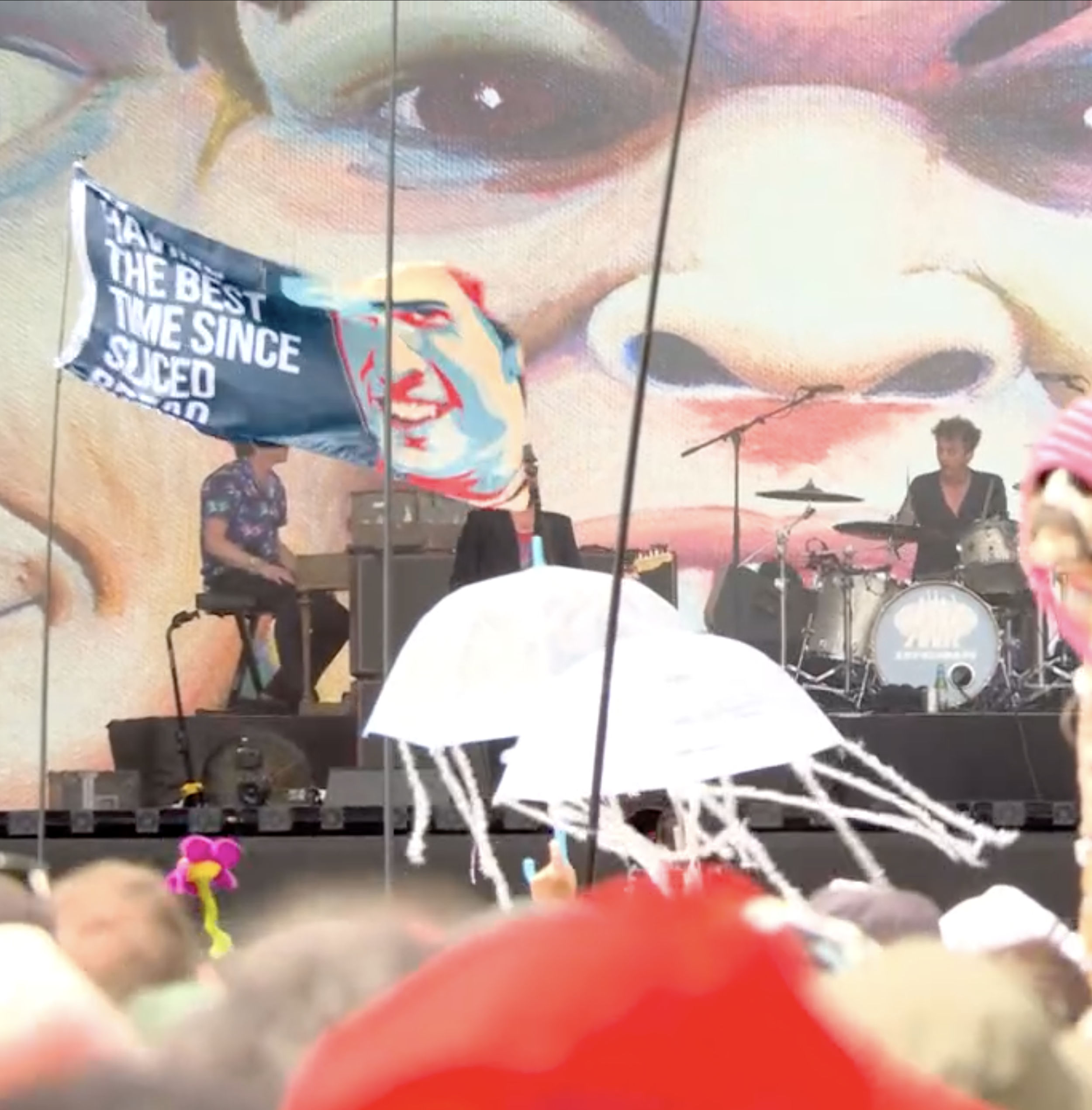 Colorful display with a large digital screen showing a woman's face, surrounded by people, some holding balloons and umbrellas, in an outdoor event setting.