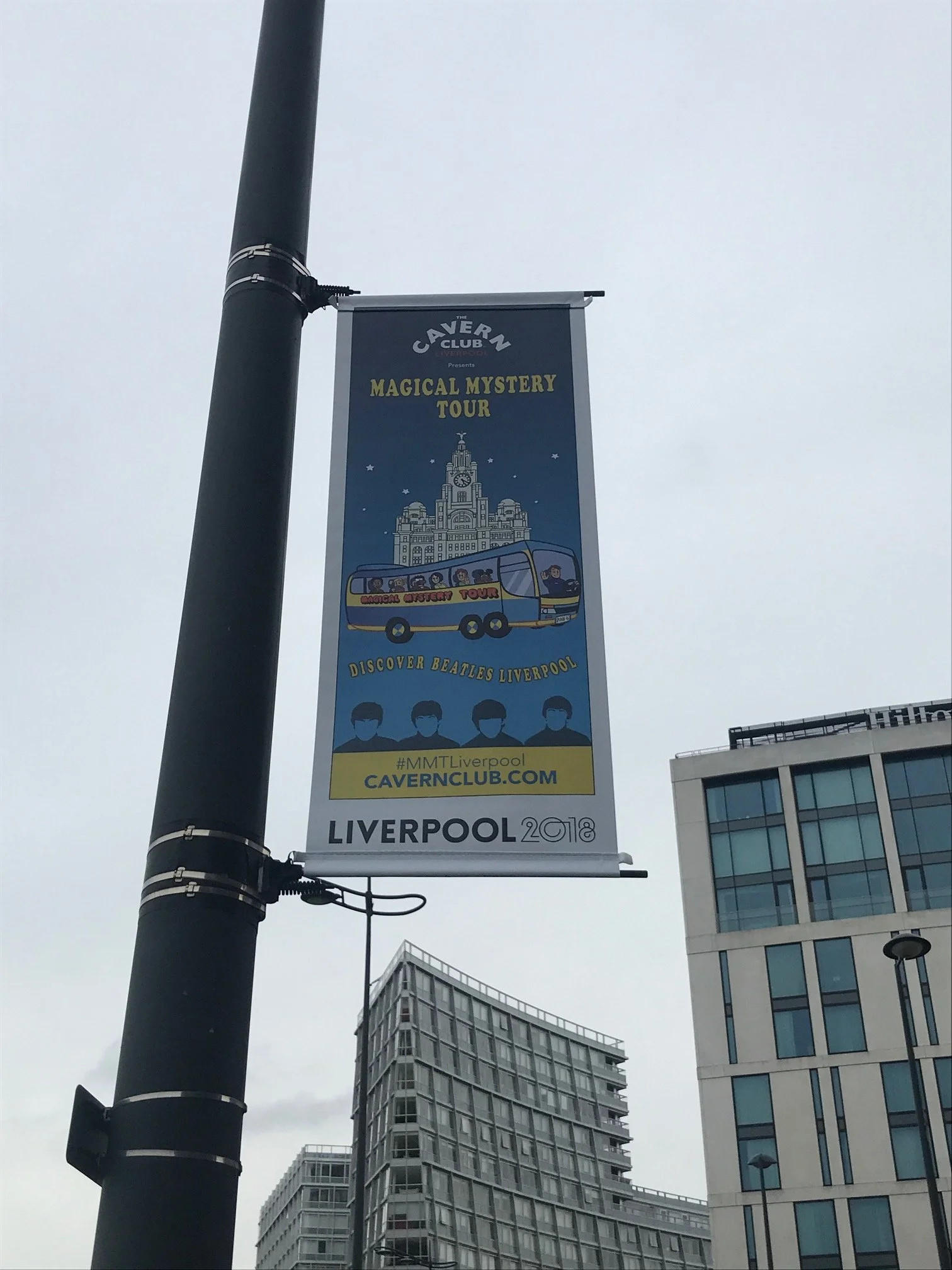 A vertical banner hanging from a black lamppost features an advertisement for the Cavern Club's Magical Mystery Tour in Liverpool, England, with illustrations of a Beatles tour bus and historic buildings, against a cityscape background with modern of