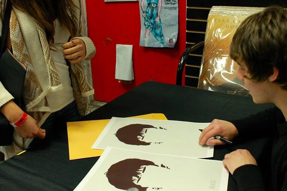 Person sitting at a signing table with multiple copies of a silhouette artwork, while a woman stands nearby in a room with red walls and posters.