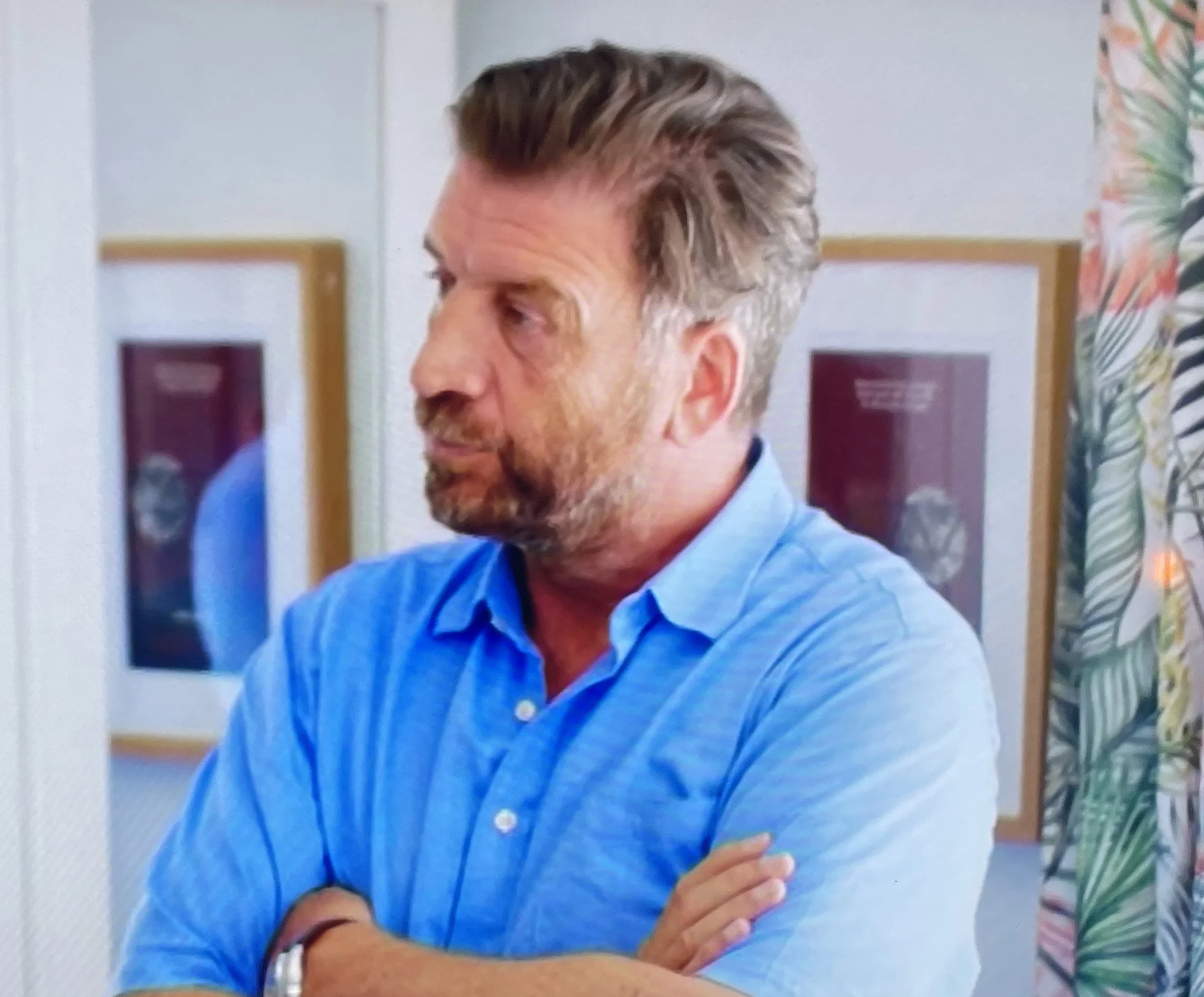 A man with brown hair and a beard wearing a blue shirt, standing indoors with his arms crossed.