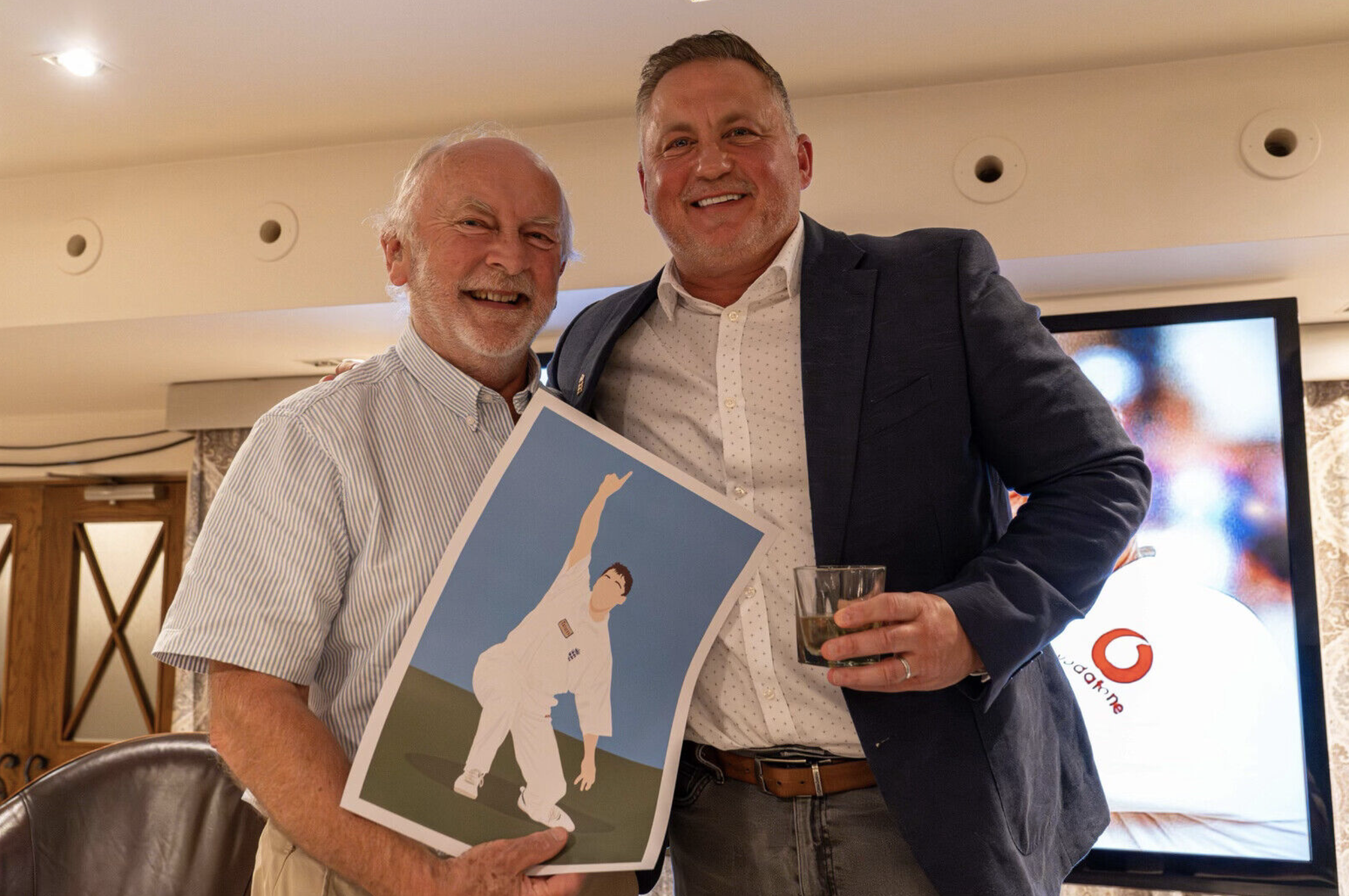 Two men smiling and standing close together at an indoor event, with one holding a framed illustration of a person in a relaxed pose, and the other holding a glass of beverage.