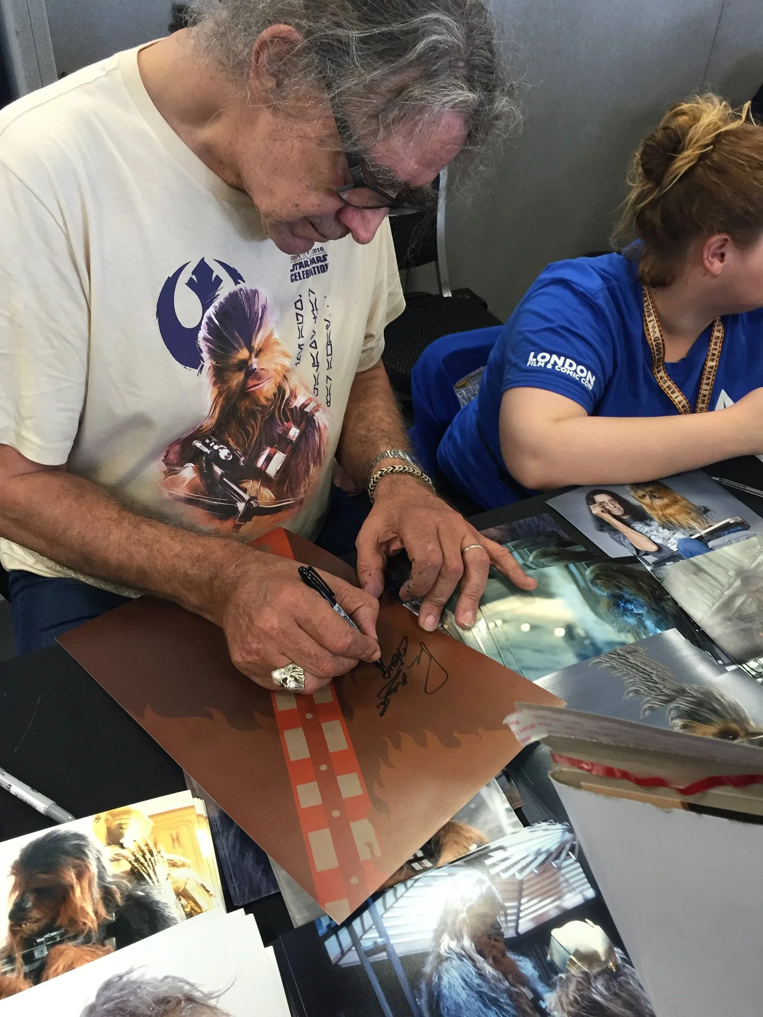 A man with gray hair and glasses signs an item for a woman who has reddish-blonde hair tied back, sitting at a table with photographs of dogs.
