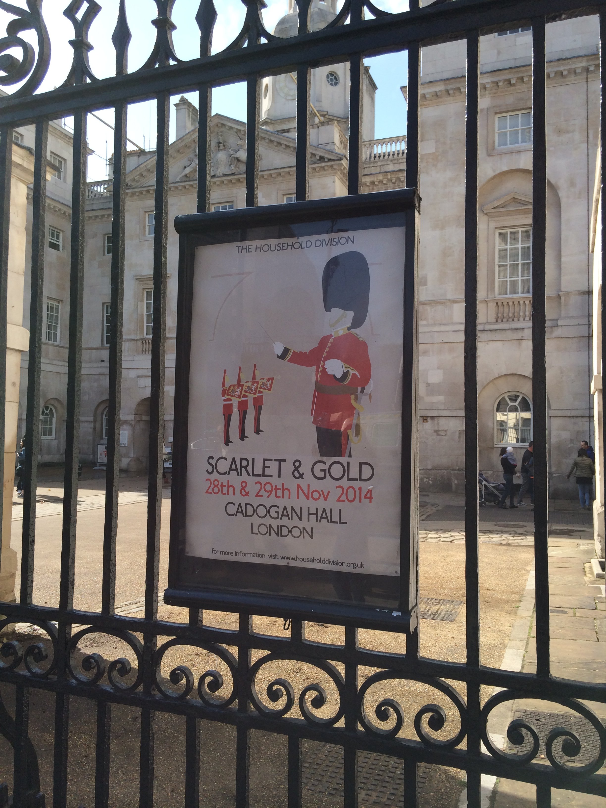 A poster behind a black iron fence advertising the Scarlet & Gold event on November 28-29, 2014, at Cadogan Hall in London, with an illustration of a british soldier and a marching band.