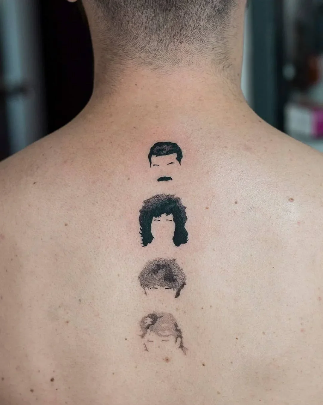 Tattoo of four minimalist black and gray portraits of famous musicians on a person's back, from top to bottom: Freddie Mercury, Brian May, Freddie Mercury again, and Kurt Cobain.
