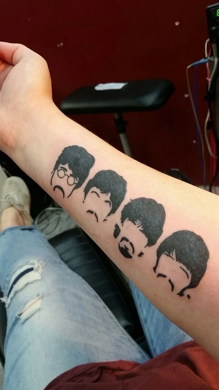 Tattoo of four black silhouettes of men's faces with distinct hairstyles and facial expressions on a person's forearm. The person is wearing ripped jeans and is seated indoors.