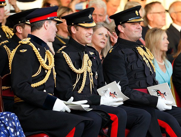 Three military officers dressed in formal black uniforms with gold braid, seated at a ceremony, holding programs, with a crowd of civilians in the background.