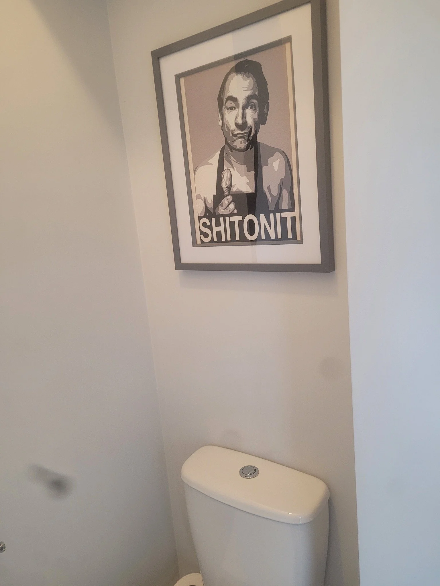 A framed black and white poster of a man with a casual expression, holding up two fingers, hanging on a white wall above a white toilet with a closed lid in a bathroom.
