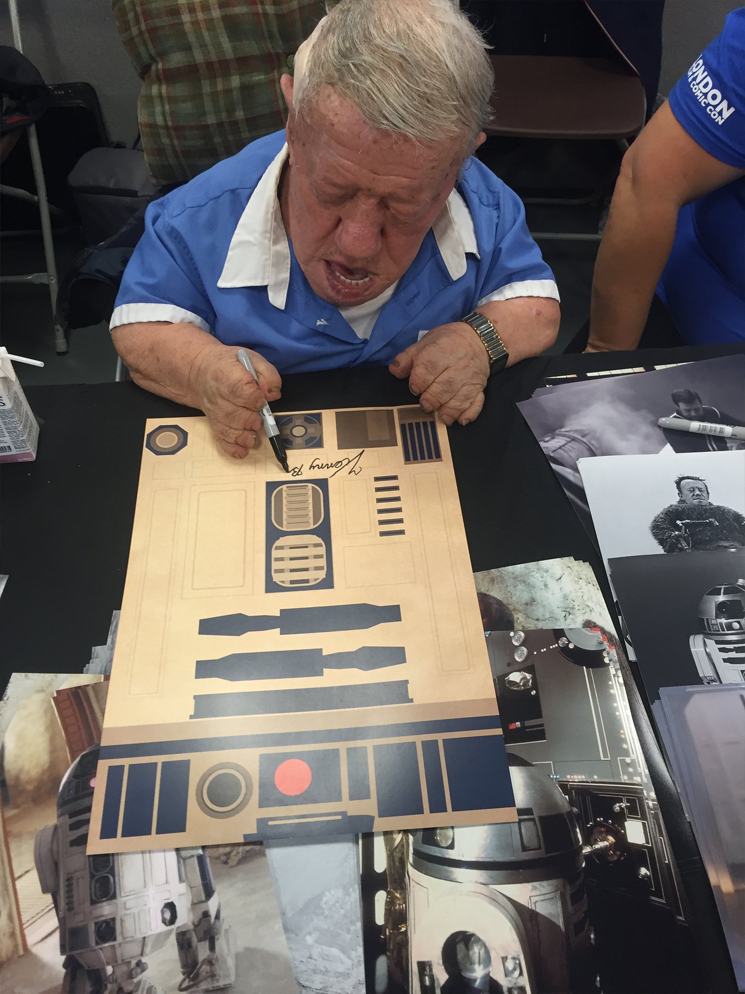 An elderly man with light hair is sitting at a table, signing a large poster or print featuring science fiction or movie robot characters, possibly R2-D2 from Star Wars, with various other black-and-white images of robots and mechanical objects aroun
