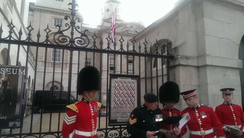 Four British soldiers in ceremonial dress, including red coats and tall bearskin hats, standing near a gate with a guard officer and a police officer reading a document, outside a historic building with the Union Jack flag.