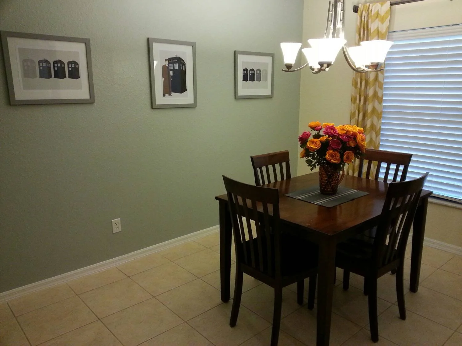 A dining room with a wooden table and five matching chairs with dark wood finish. On the table is a large vase filled with pink, orange, and yellow flowers. The wall behind has three framed pictures of vintage-style cartoon-like images of the TARDIS 