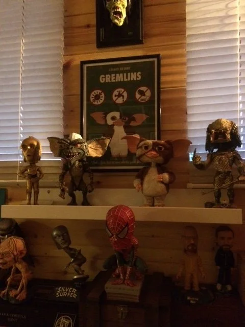 Shelf displaying various collectible figurines, including a small skull, a creature with sharp teeth, a plush cat, and a character with glasses. Behind the shelf, there are framed posters of Gremlins and other themed artwork, with wooden paneling and