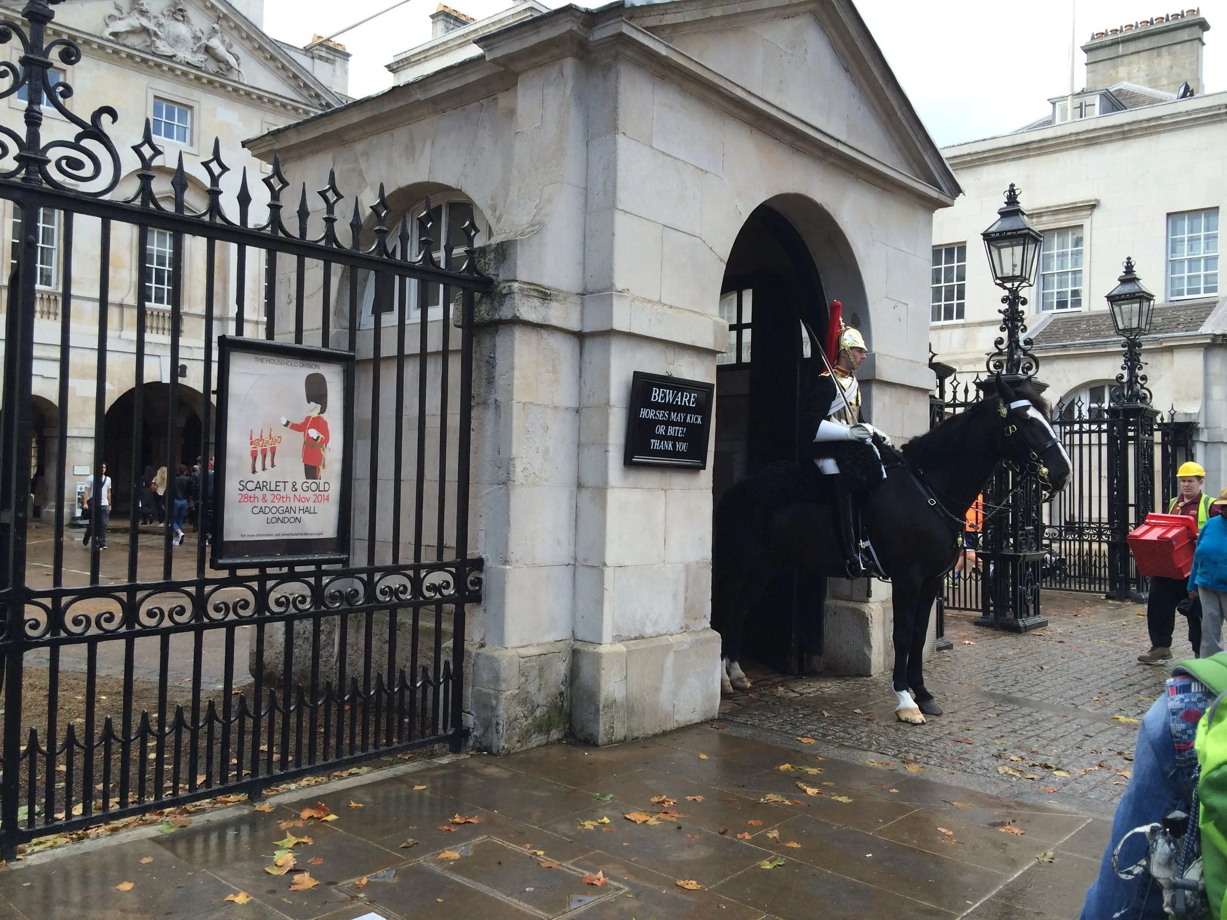 A guard dressed in traditional historical attire, riding a black horse in front of a stone building with iron gates. Several people are walking nearby in an urban setting, with signs on the gate warning about horses and a poster advertising an exhibi