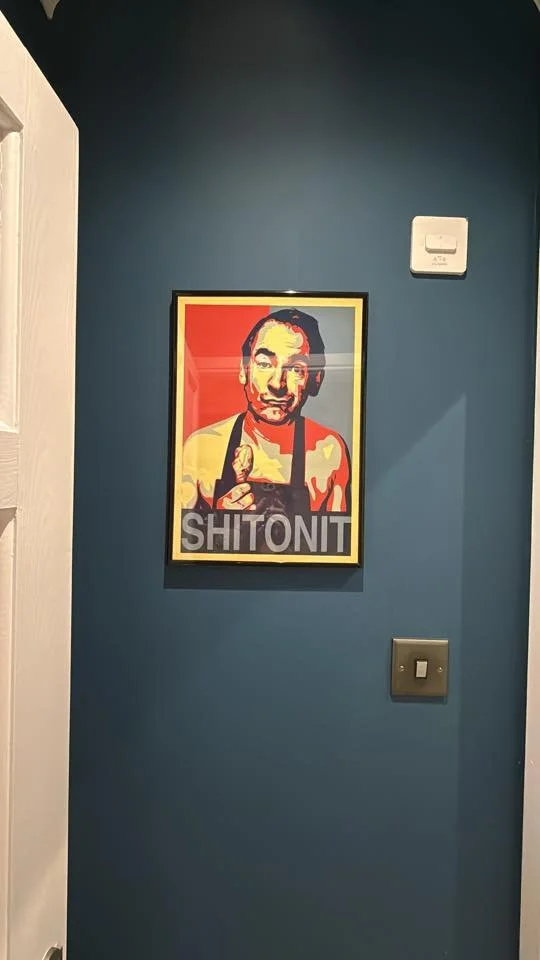 A framed pop art poster on a dark blue wall depicts a man with dark hair, pointing one finger, with the word "SHITONIT" written below in bold letters.