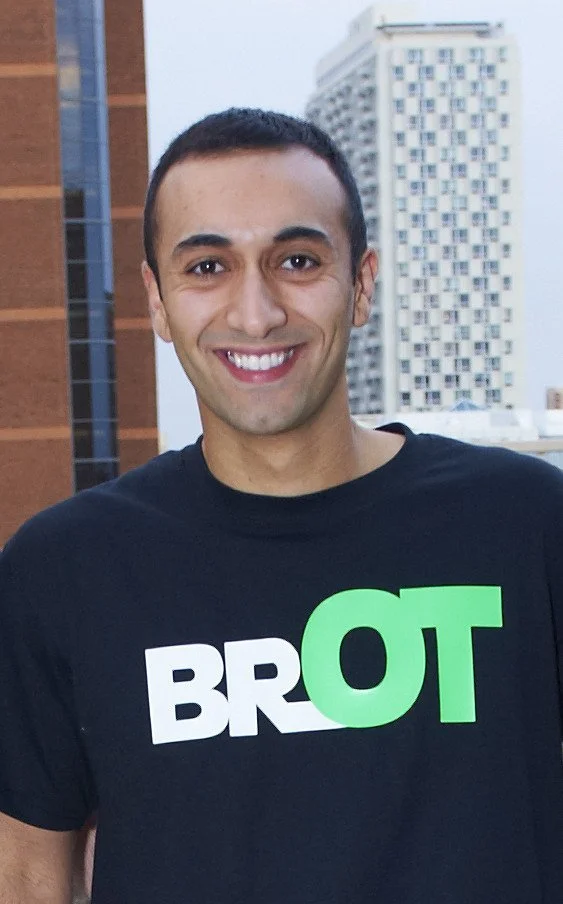 Smiling man with short dark hair, wearing a black T-shirt with bold white and green text, standing outdoors in an urban setting with tall buildings in the background.