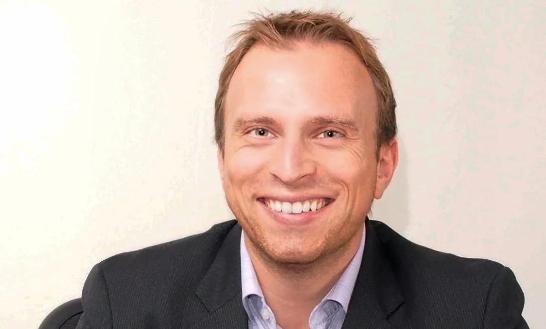 Headshot of a smiling man in a suit sitting in front of a plain light-colored wall.