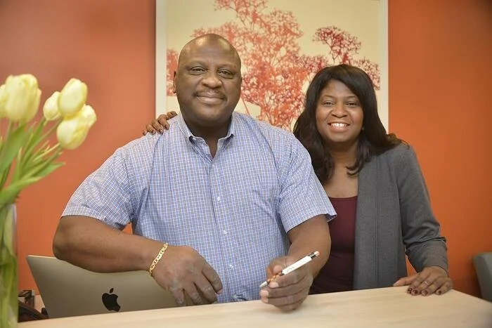 A man and woman smiling and sitting at a table in an office, with a vase of tulips on the left and a framed picture of trees on the wall behind them.