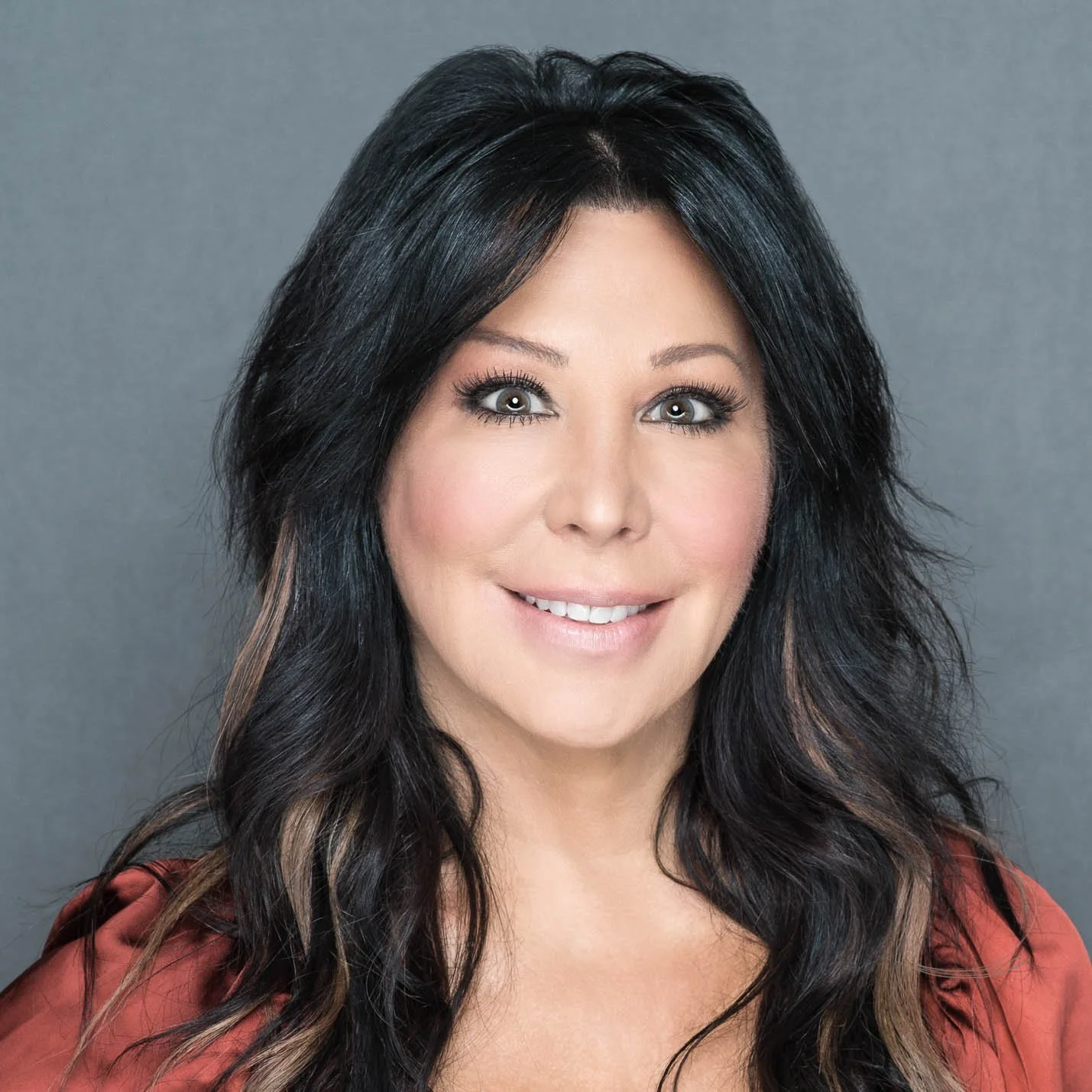 A woman with long dark hair, styled with loose waves, smiling against a gray background.