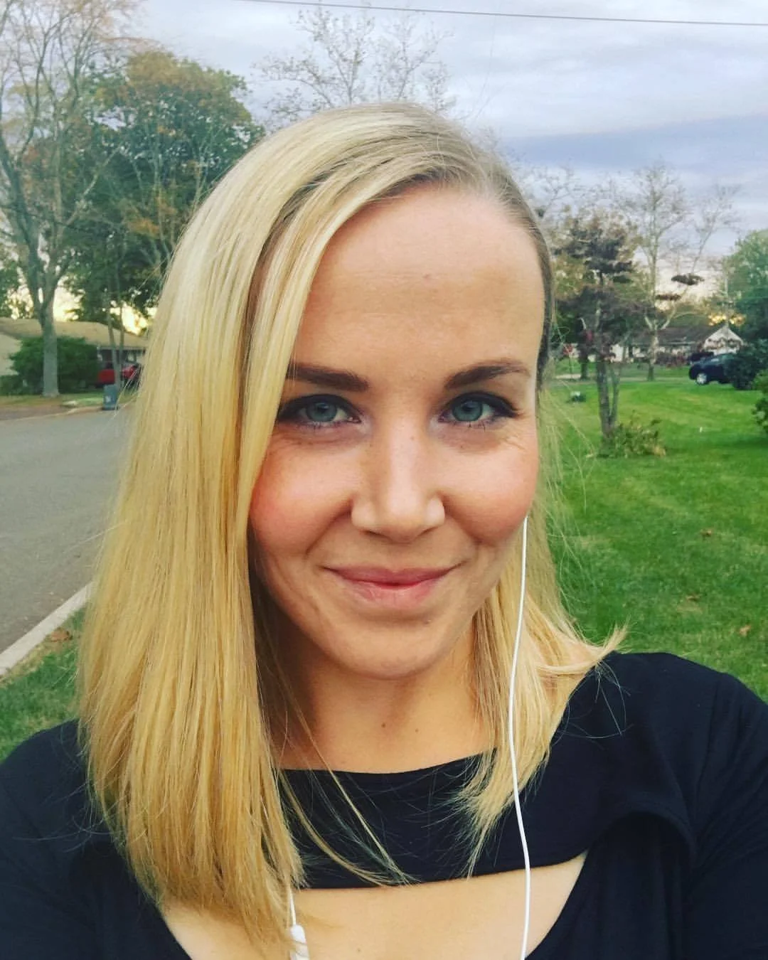 Close-up outdoor selfie of a woman with blonde hair and blue eyes, smiling, wearing a black top and earphones, in a green neighborhood park or yard during daytime.