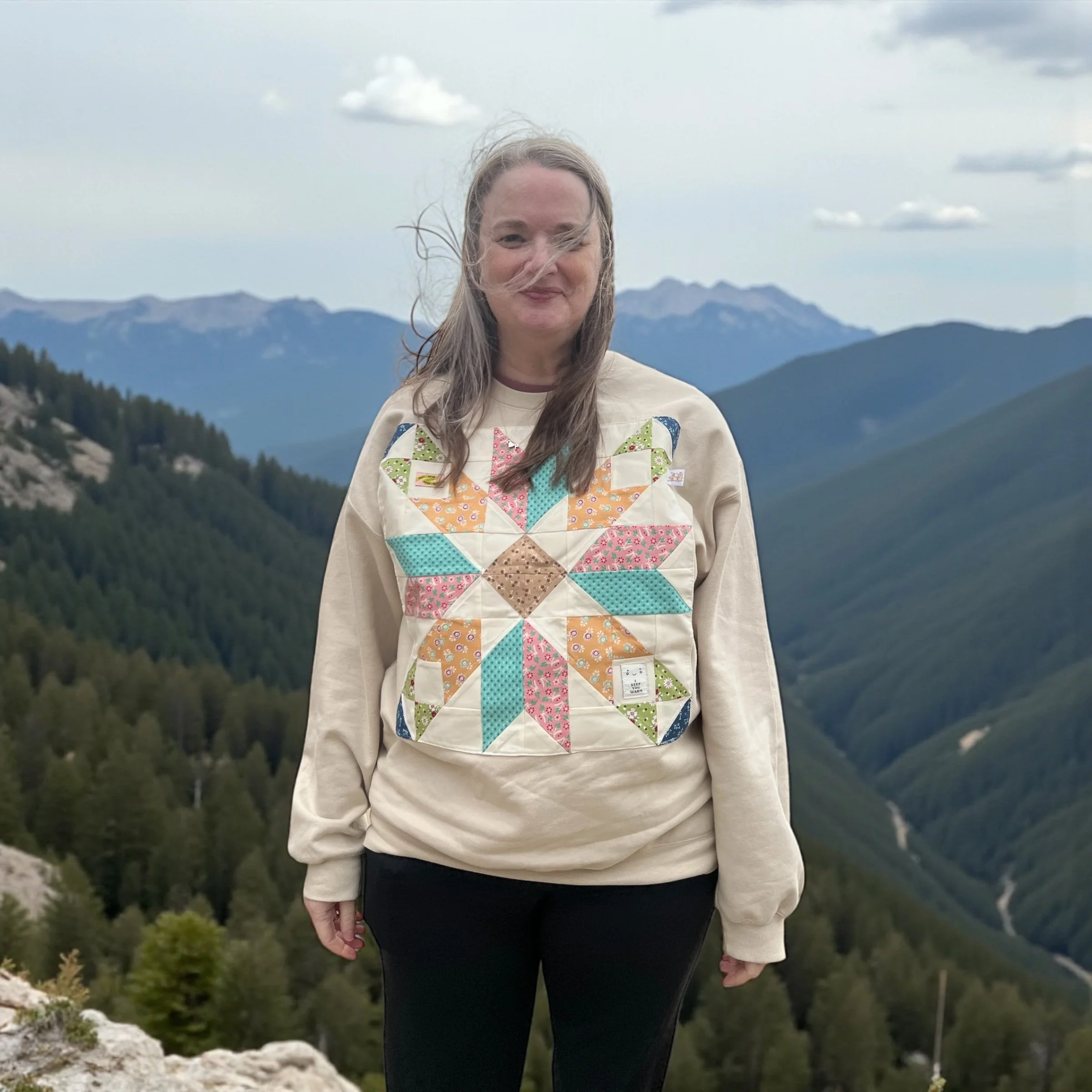 A woman standing outdoors on a mountain trail with forested mountains in the background, wearing a beige sweatshirt with a colorful quilted star design on the front, and black pants.
