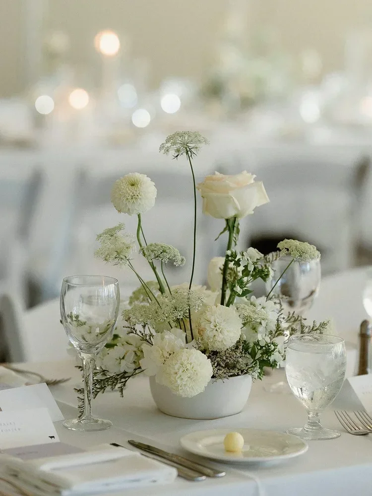 Elegant table setting with a centerpiece of white flowers including roses, dahlias, and Queen Anne's lace, glassware, a plate with a butter pat, and silverware on a white tablecloth.
