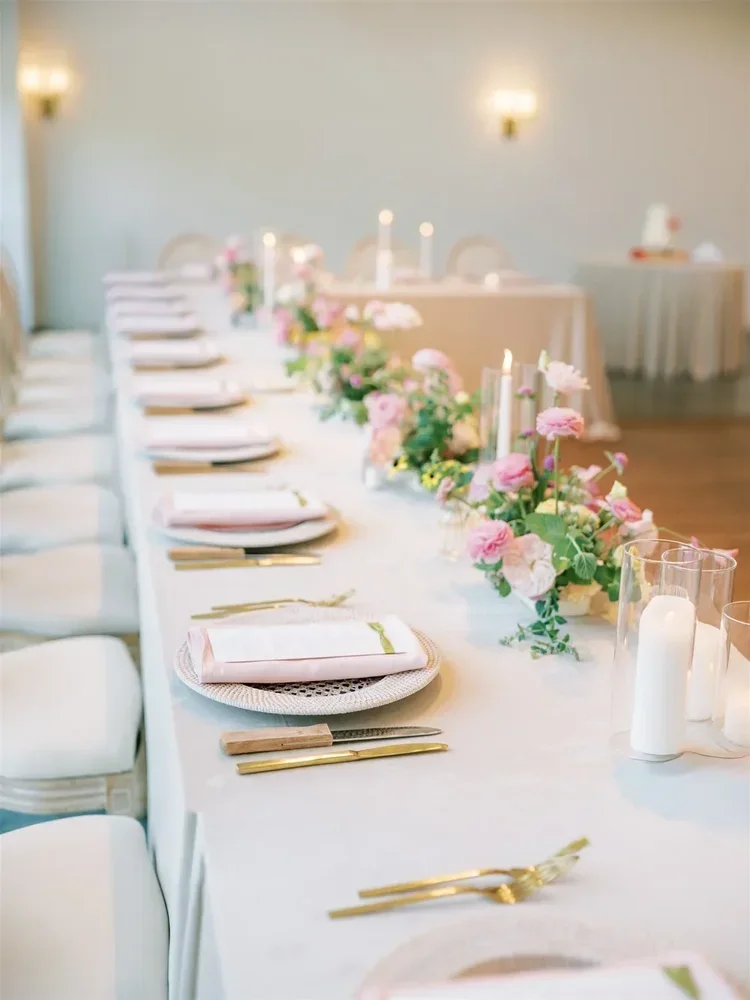 A long dining table set for a formal event with pink and white floral centerpieces, candles, plates, napkins, and gold flatware.