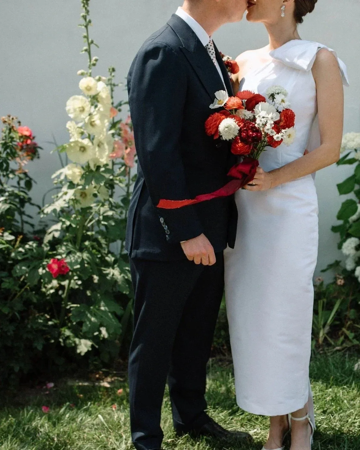 A couple dressed in wedding attire sharing a kiss while holding a bouquet of red and white flowers, standing outside with white and pink flowers in the background.