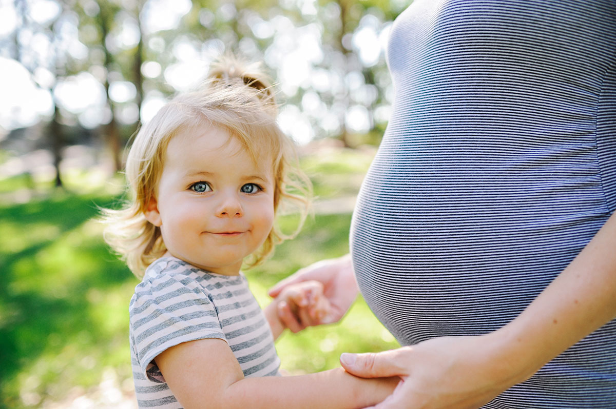 Toddler holding mum's hands with her face near mum's pregnant belly