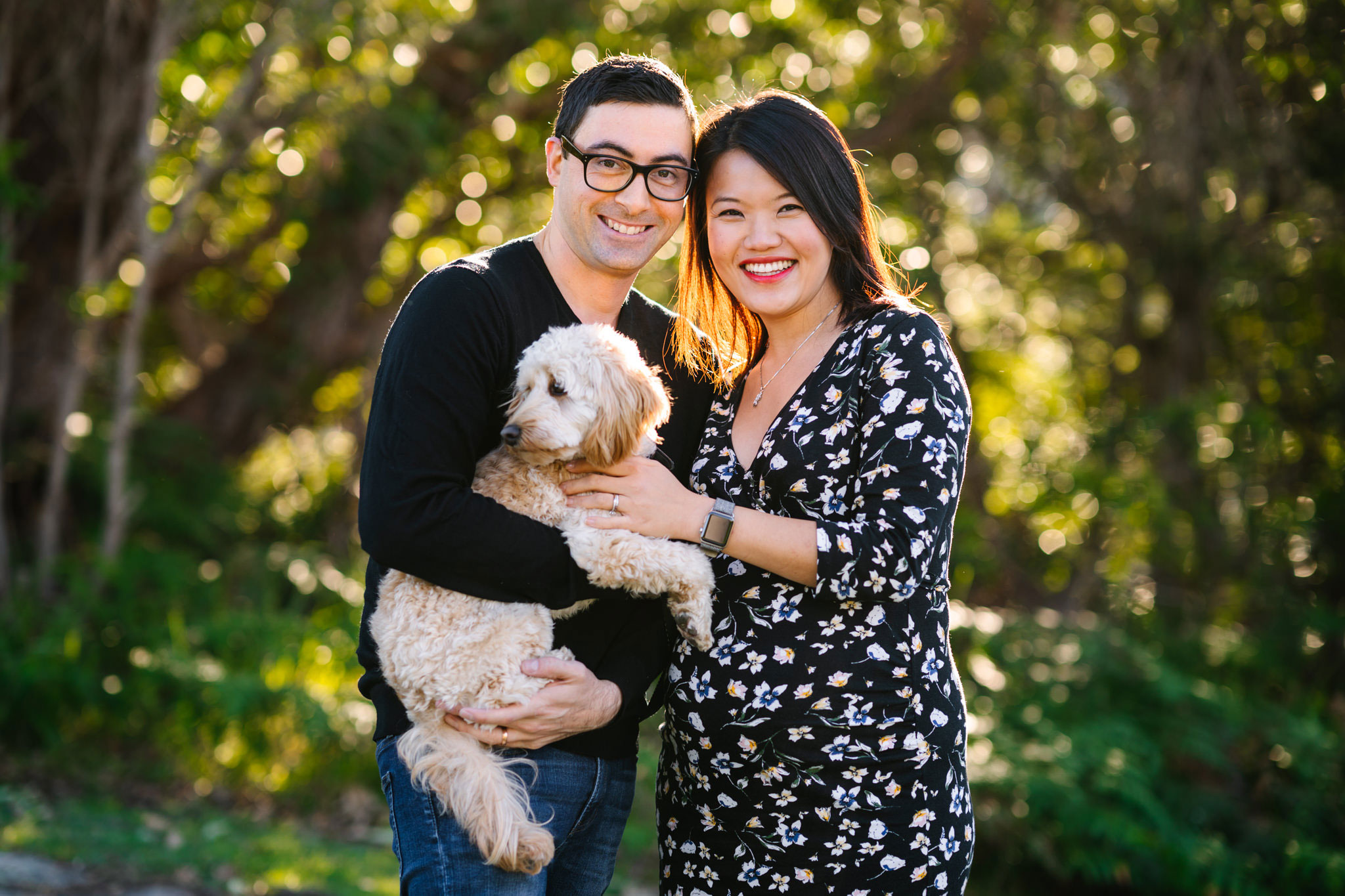 Couple holding their beige poodle with trees in the background