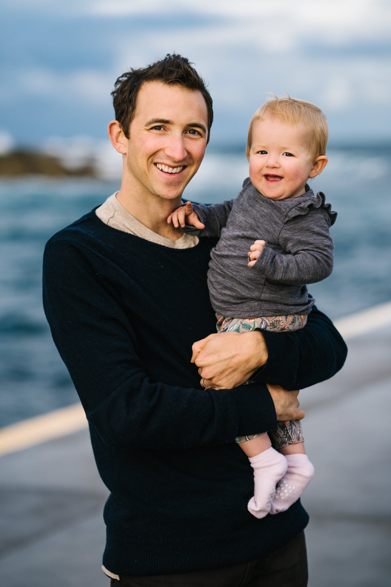 Baby and dad smiling at the camera with beach in the background