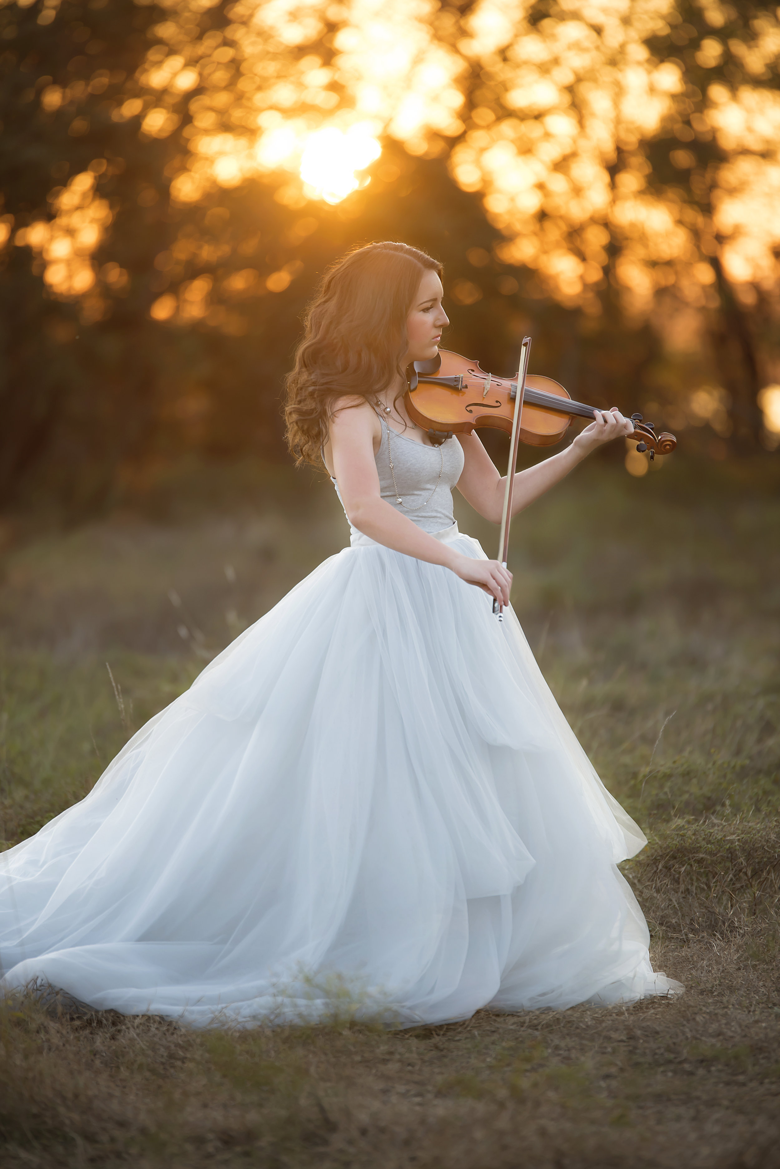  Woman in white dress playing violin in a green field at sunset. 