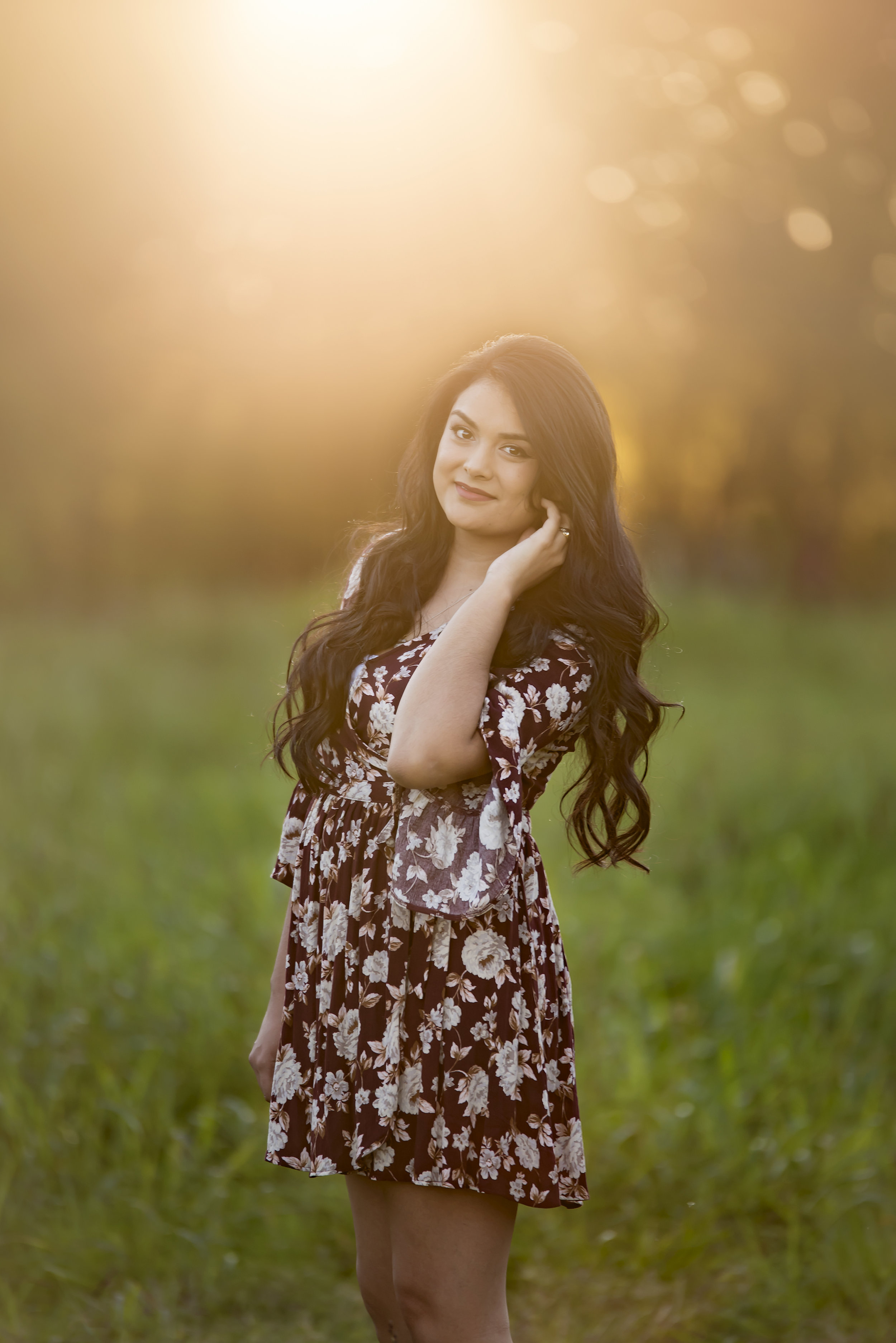  Senior heirloom portrait at sunset, girl standing in a field in a floral dress, facing the camera. 