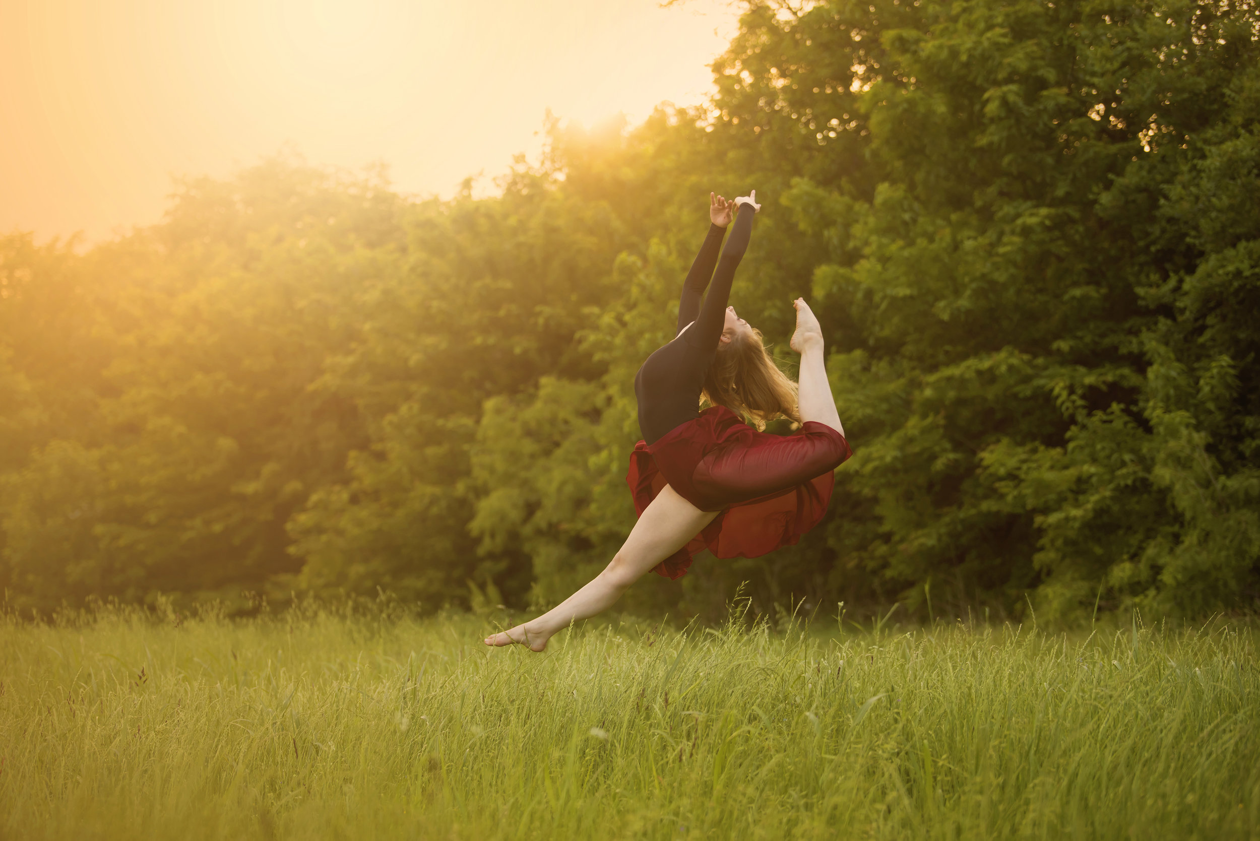  Sunset over trees in the background, woman in red dress jumping with legs and arms outstretched. 