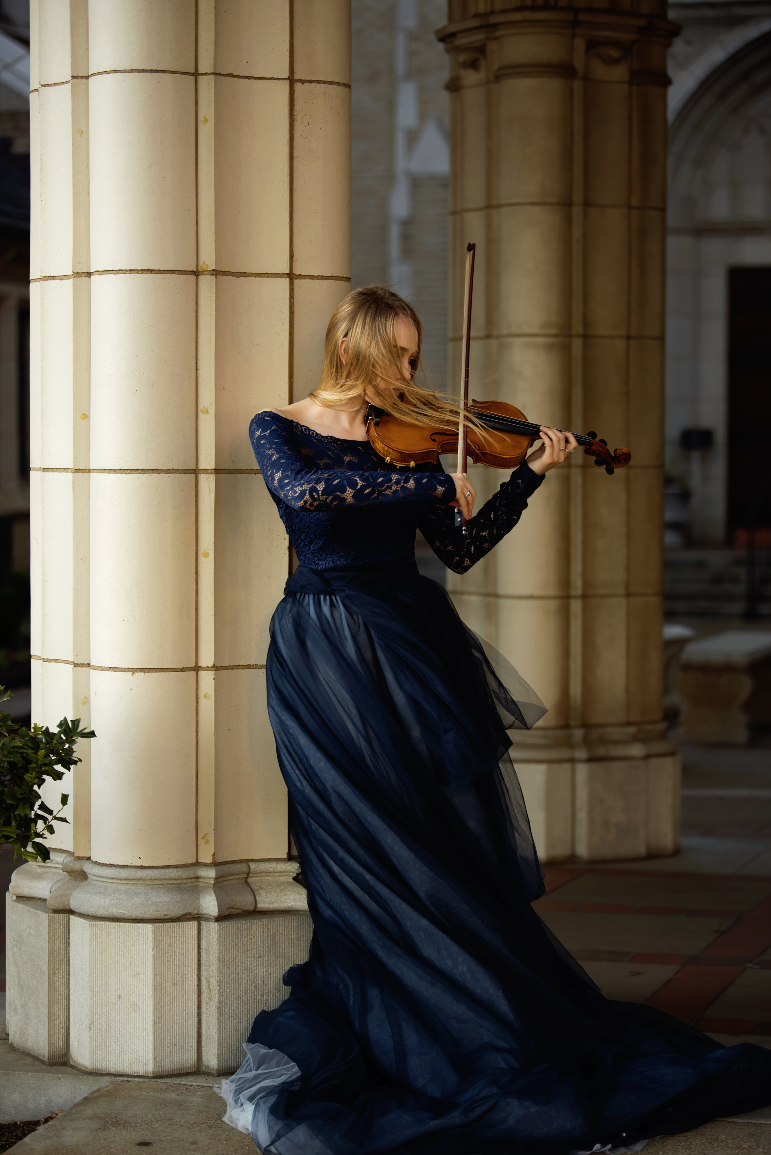  Active portrait, woman in blue dress paying violin standing next to columns. 
