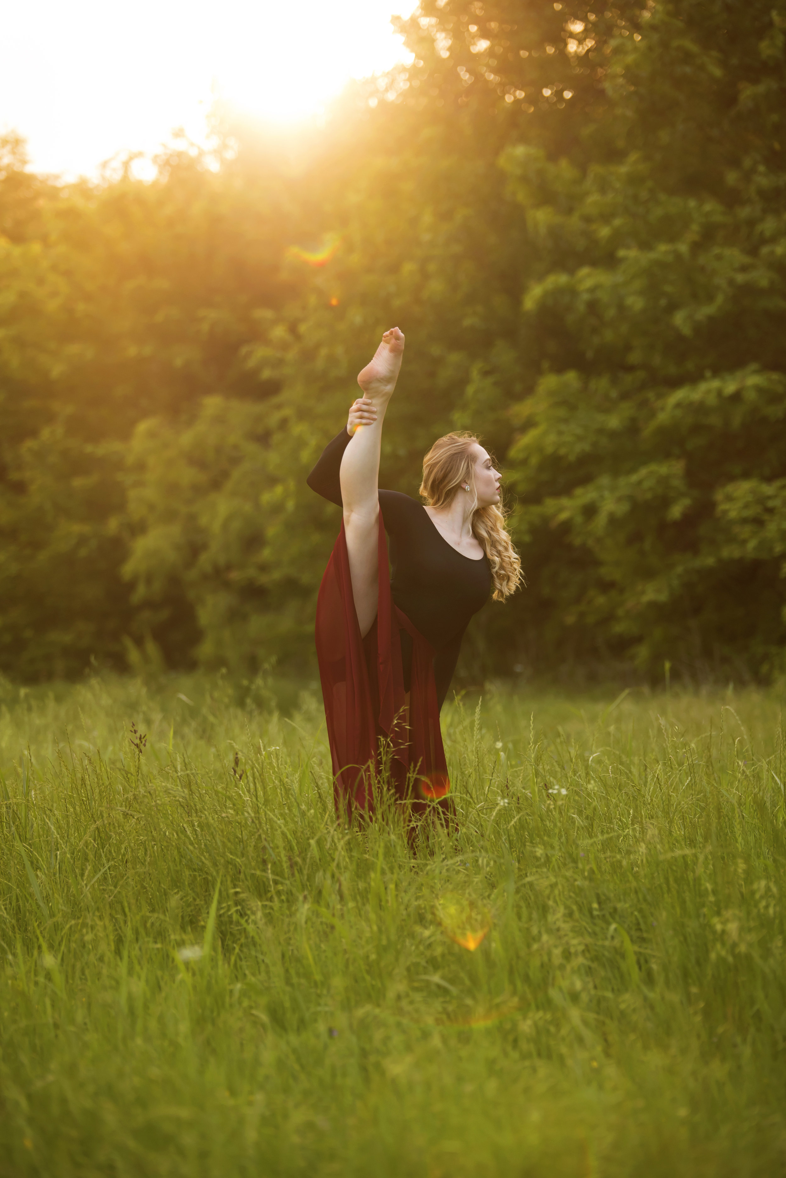  Lifestyle portrait of woman in field at sunset, looking away from camera while holding one leg extended in the splits. 