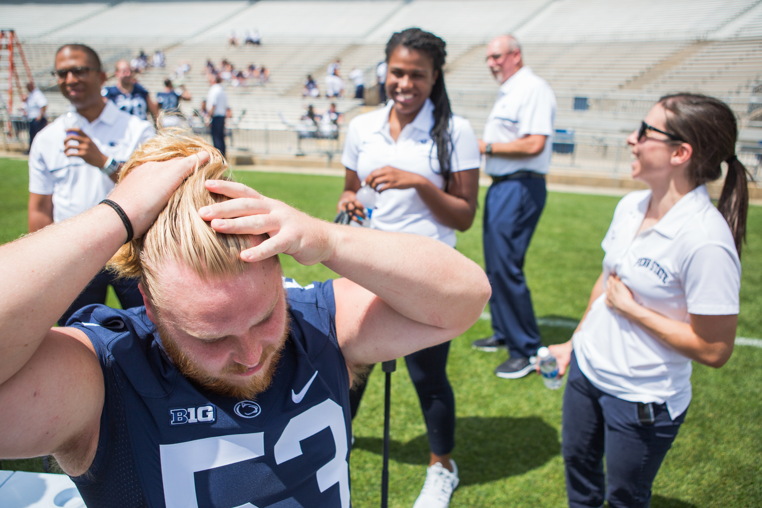 Graduate student Derek Dowrey shows assistant learning specialist Chelsea Holmes, center, and assistant athletic trainer Madeleine Scaramuzzo, right, how he ties his hair into a bun during the football team's photo day on the field of Beaver Stadium on Sunday, Aug. 7, 2016.