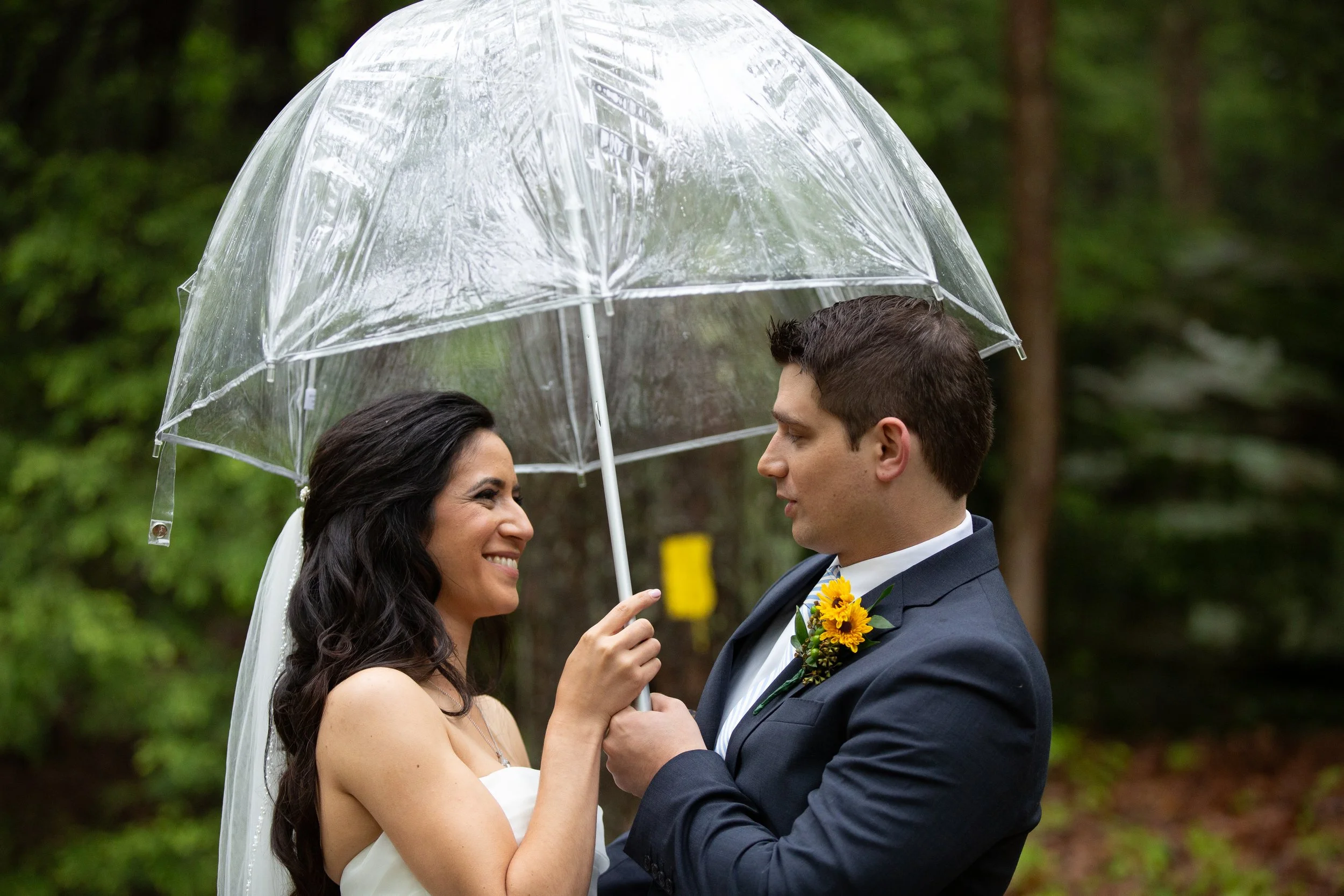 A bride and groom standing close together under a transparent umbrella during an outdoor event, with trees in the background.