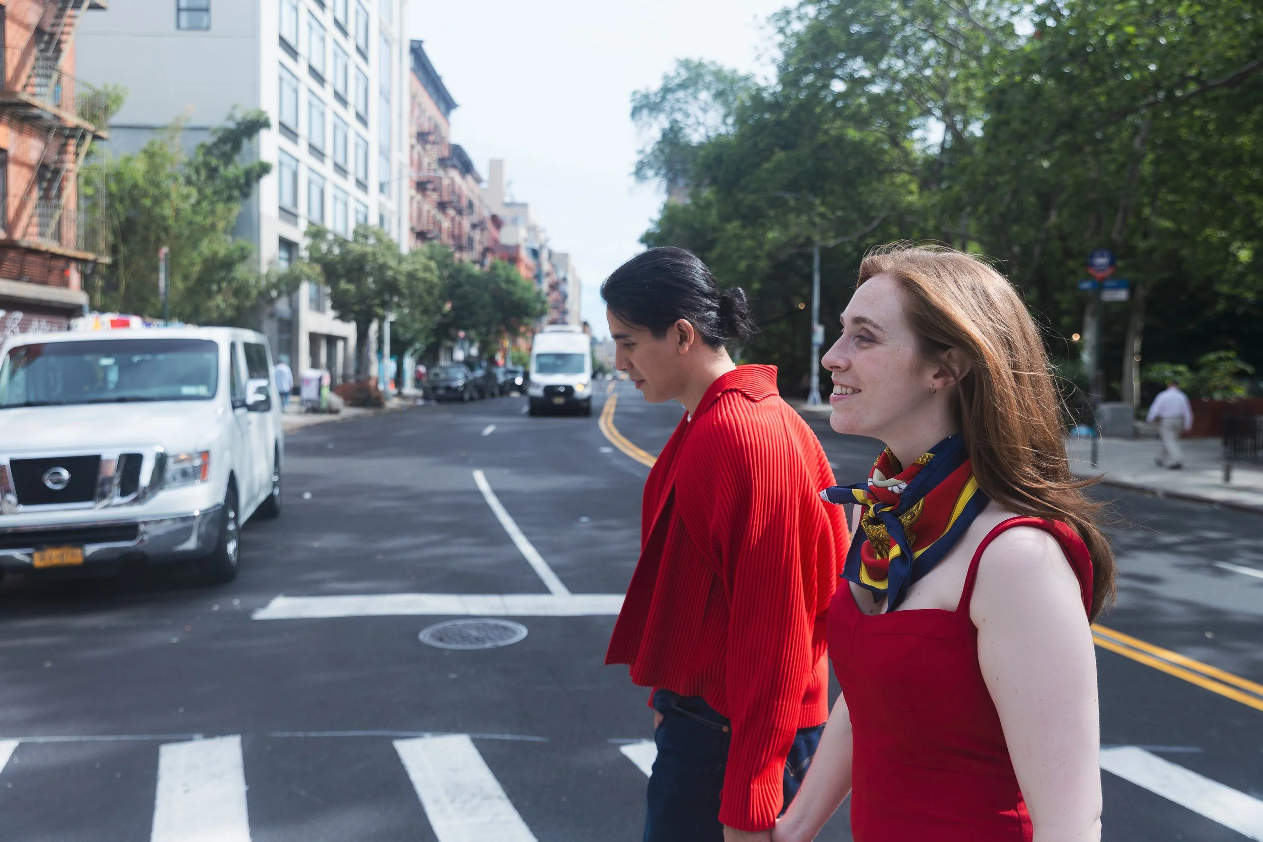 A couple walking across a city street with cars and buildings in the background. The woman in front has long red hair and is wearing a red sleeveless top with a colorful scarf. The woman beside her has short dark hair and is wearing a red jacket.