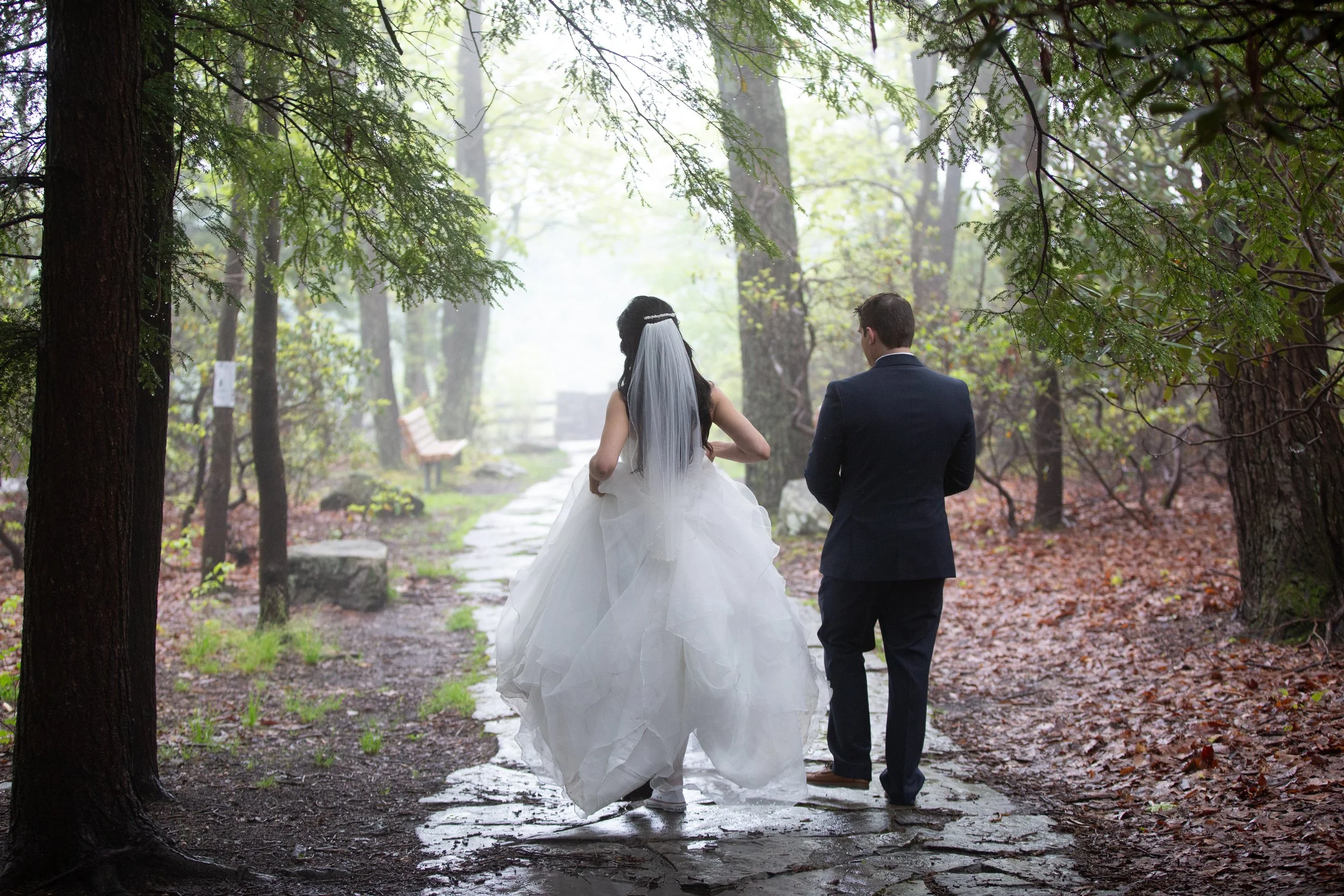 A bride and groom walk on a misty forest trail, with the bride wearing a white wedding gown and veil, and the groom in a dark suit, surrounded by tall trees and lush greenery.