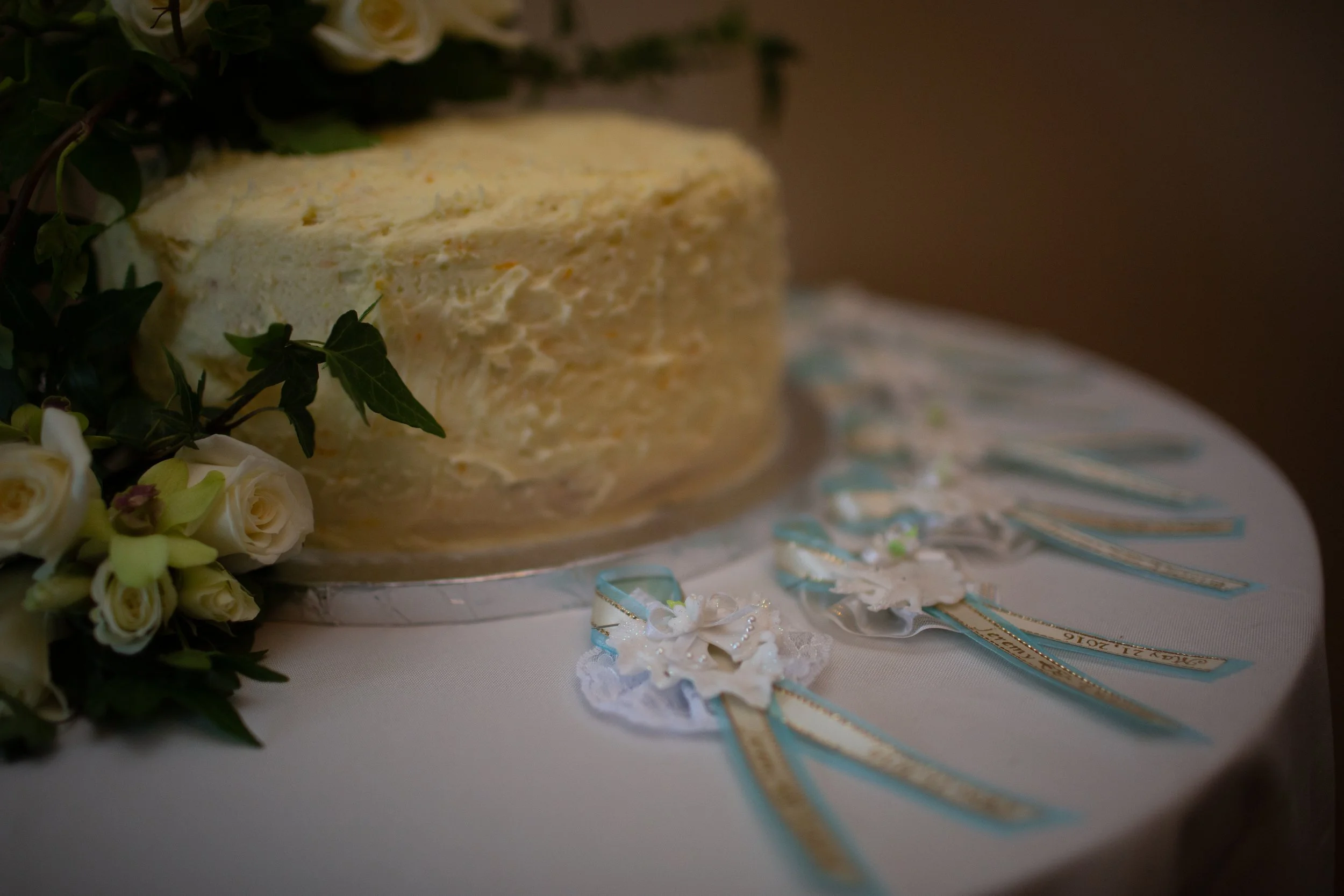 A wedding cake decorated with white frosting and surrounded by white roses and green leaves. The cake is placed on a table with pastel-colored ribbons and lace decorations.