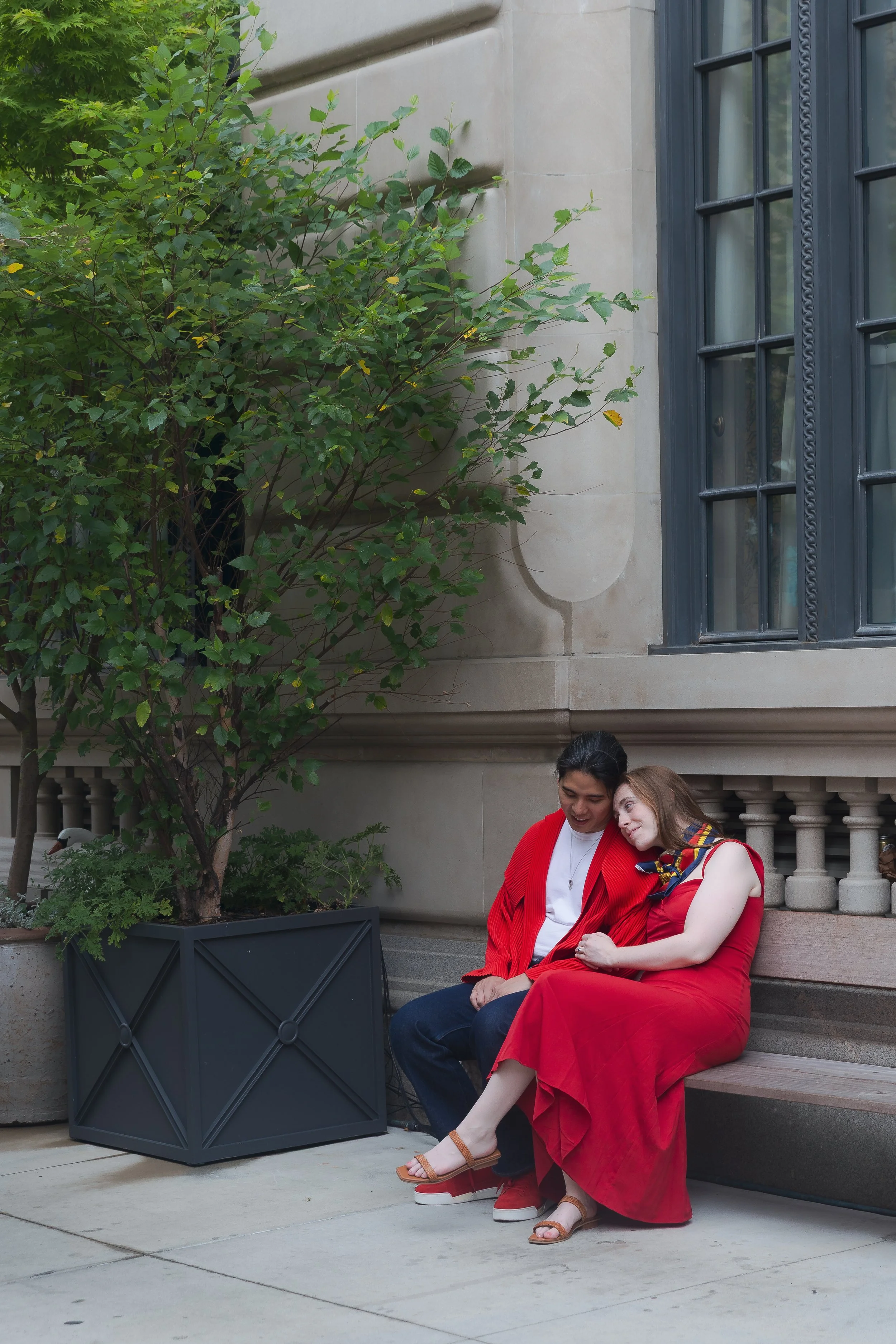 A couple sitting on a bench, one dressed in red with beige sandals, the other in dark jeans and a red jacket, near a building with large windows and a potted tree.