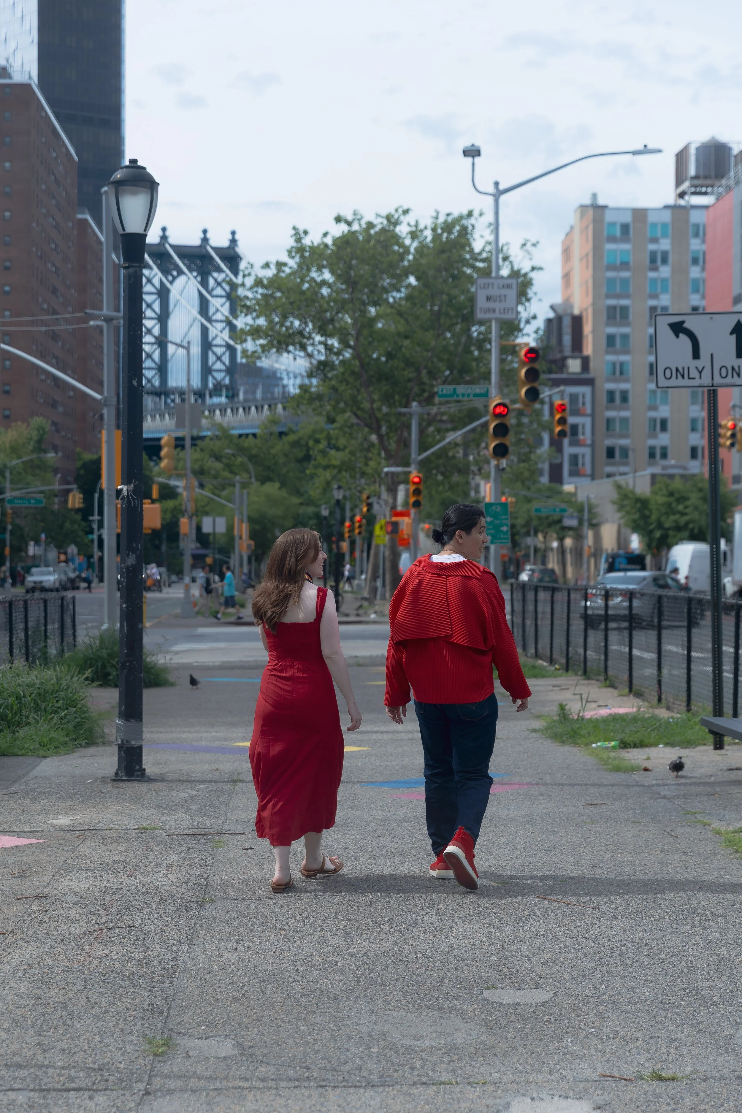 A couple walking together on a city sidewalk with tall buildings, street signs, traffic lights, trees, and cars in the background.