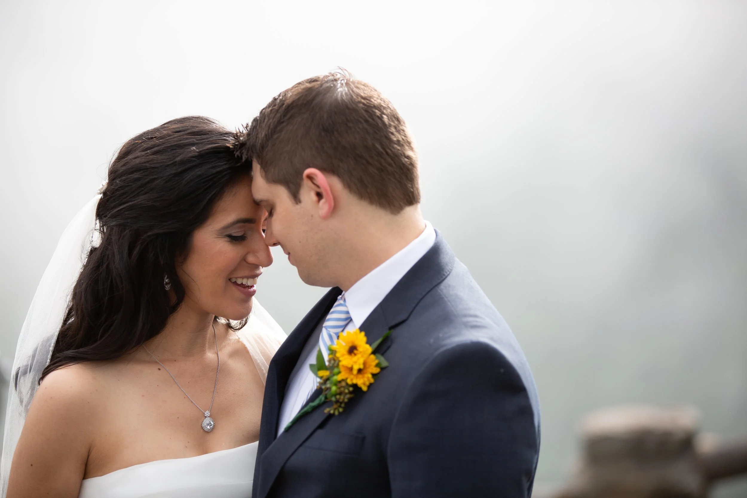 A bride and groom face each other with foreheads touching, smiling, outdoors on their wedding day, with a gray sky in the background.