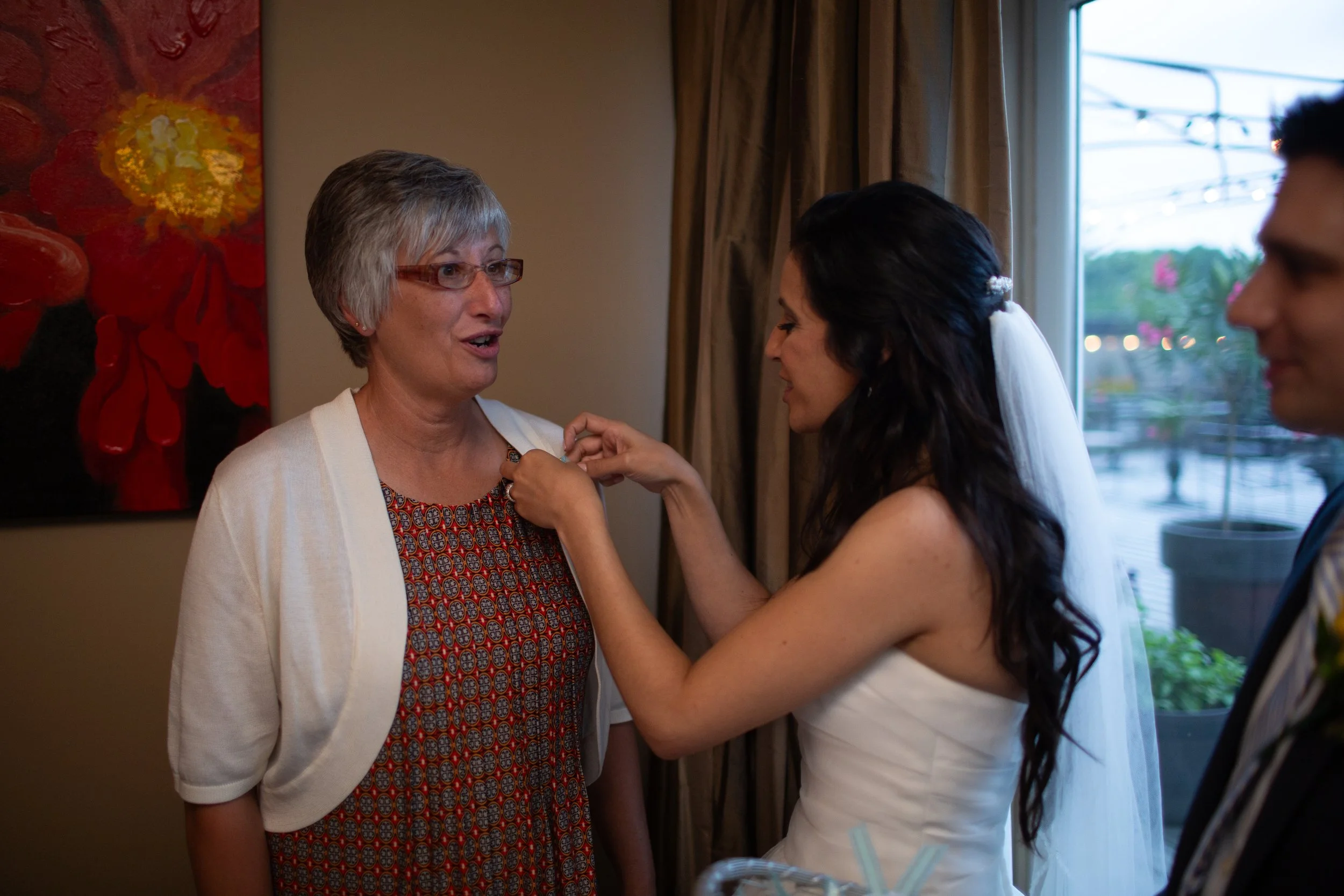 A woman with dark hair and a white dress receives a pin from an older woman with gray hair, glasses, and a patterned dress during a celebration or event inside a room with beige walls, curtains, and a window showing an outdoor view with pink flowers.