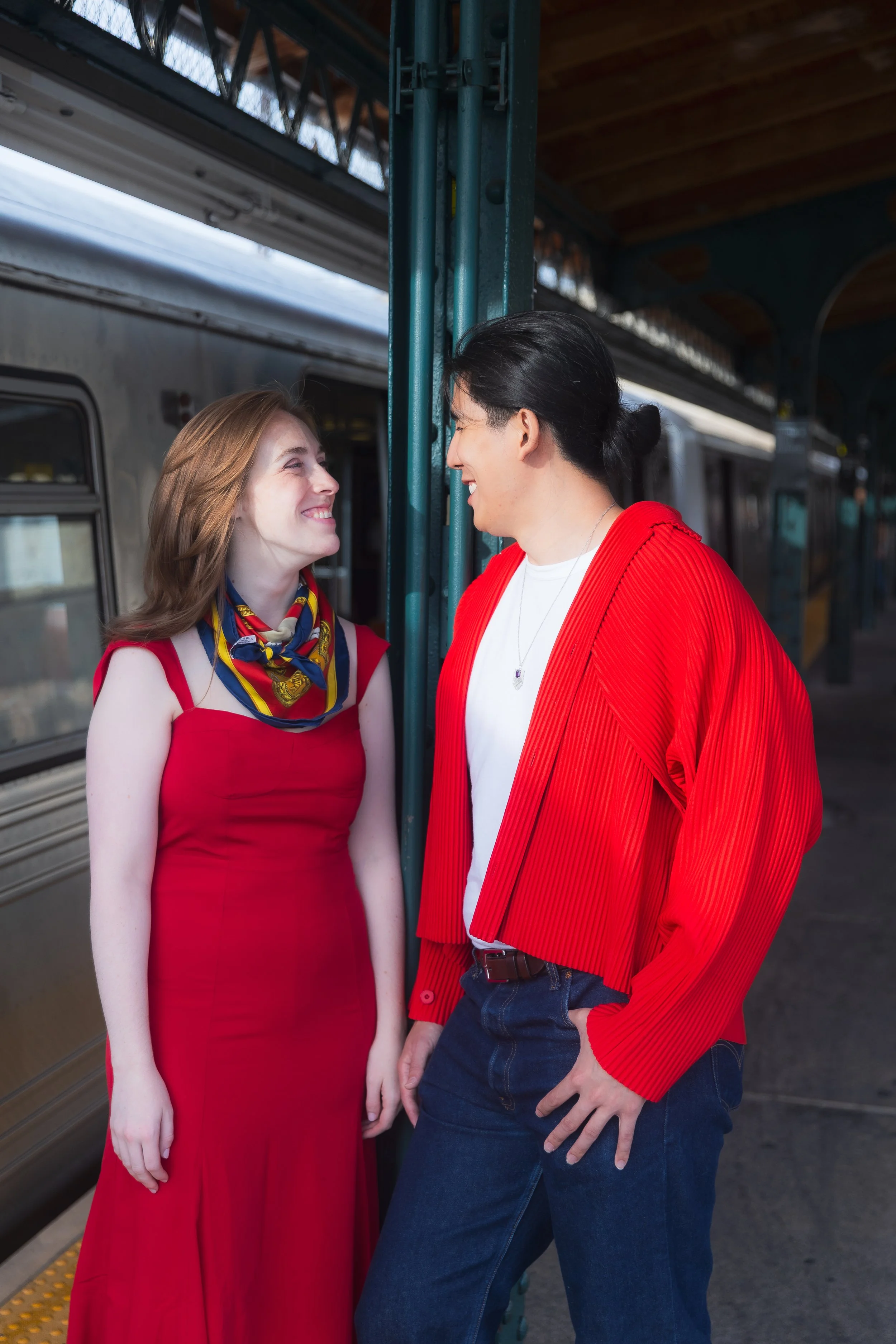 A couple smiling and facing each other at a train station platform, one wearing a red dress with a colorful scarf and the other in a red jacket and blue jeans.