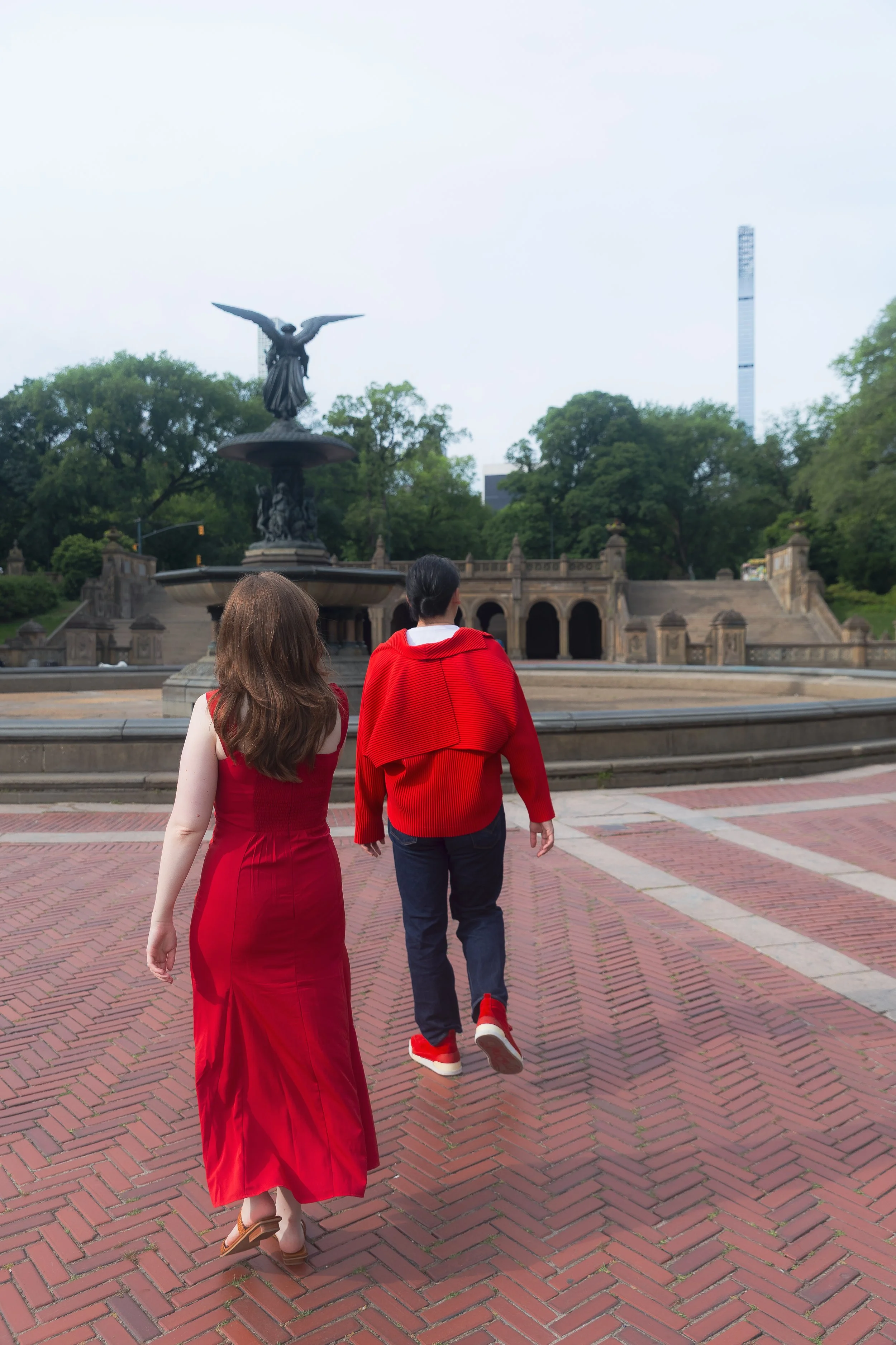Two people walk together on a brick-paved area near a fountain with a statue of an angel with outstretched wings, surrounded by green trees and a tall modern tower in the background.