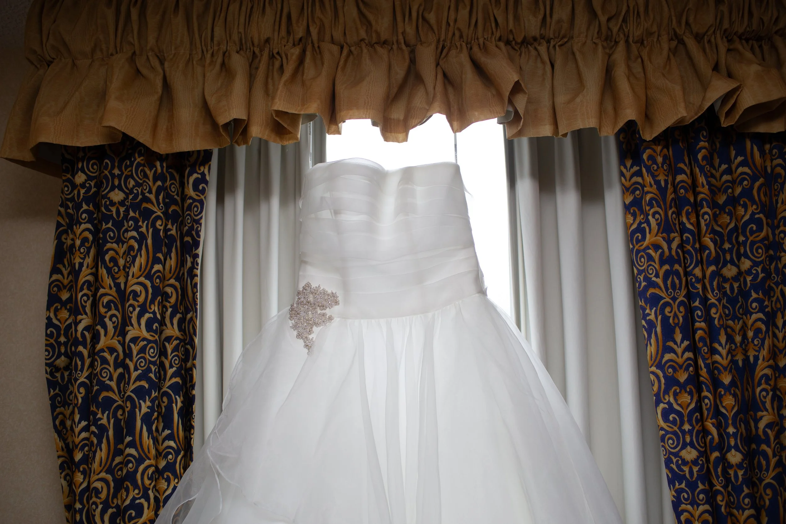 Wedding dress hanging in front of a window with patterned curtains and a valance.