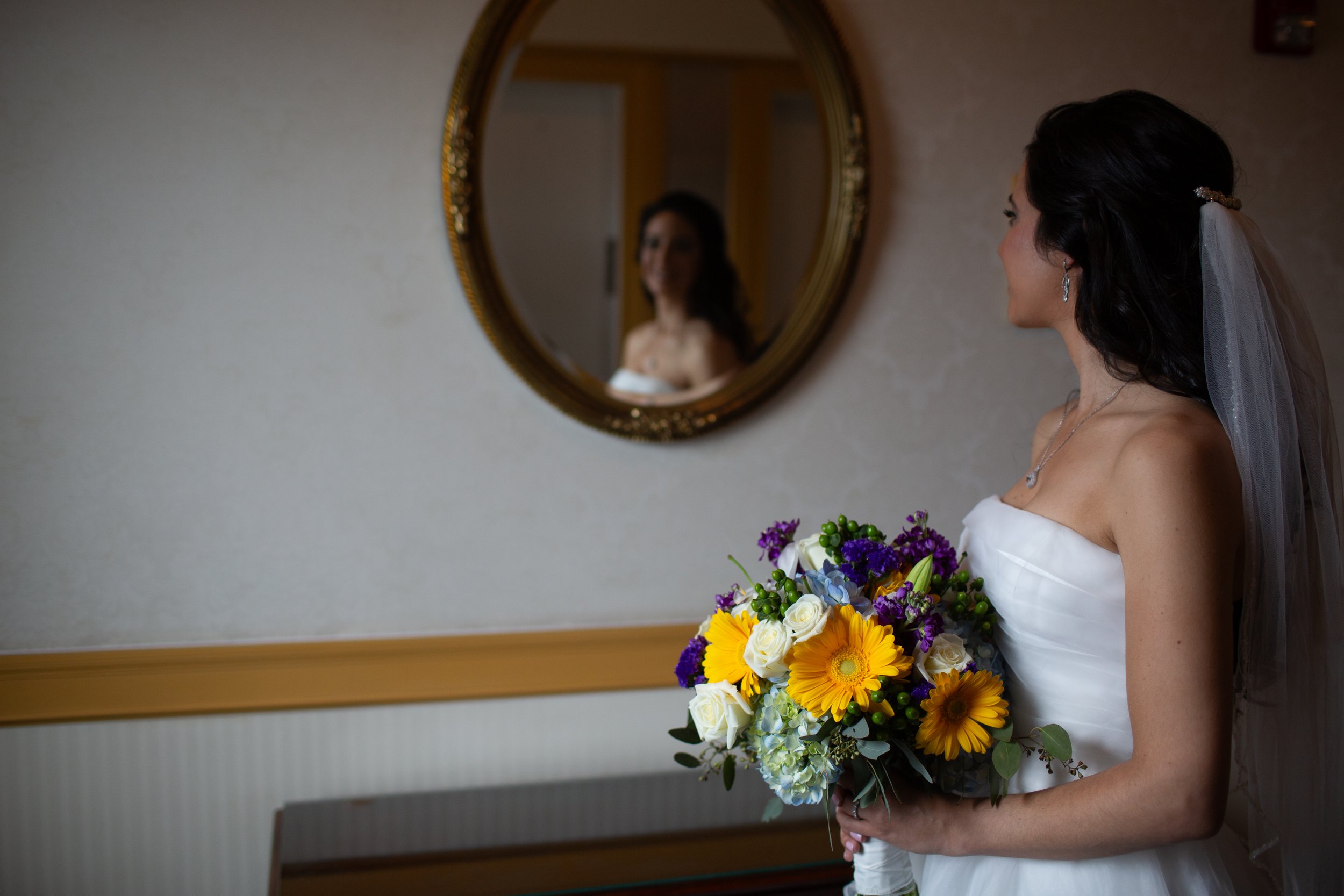 A bride with dark hair and earrings holding a colorful bouquet of flowers, looking at her reflection in a mirror on the wall.