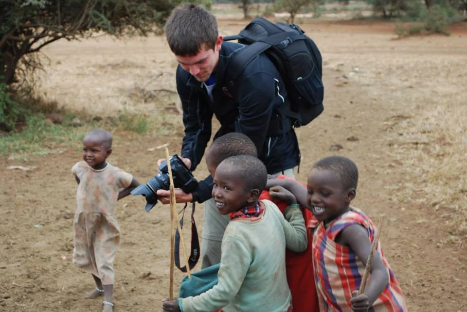 A man and four children outdoors in a dry, rural area. The man is holding a camera and appears to be engaging with the children, who are smiling and holding sticks.