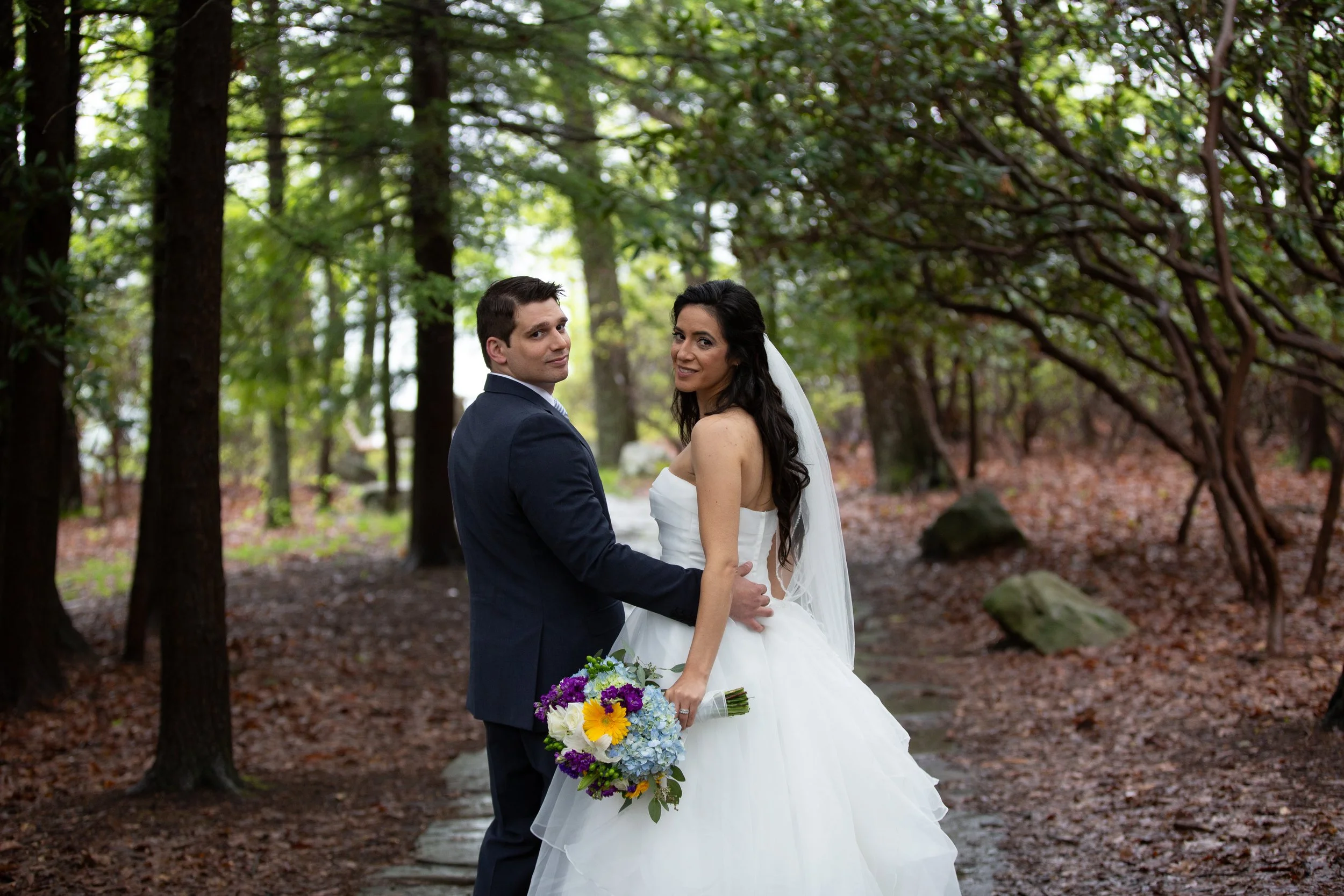 A bride and groom standing together outdoors in a wooded area during a wedding. The bride is holding a colorful bouquet and wearing a white wedding dress, while the groom is in a dark suit. They are looking at the camera with trees and fallen leaves 